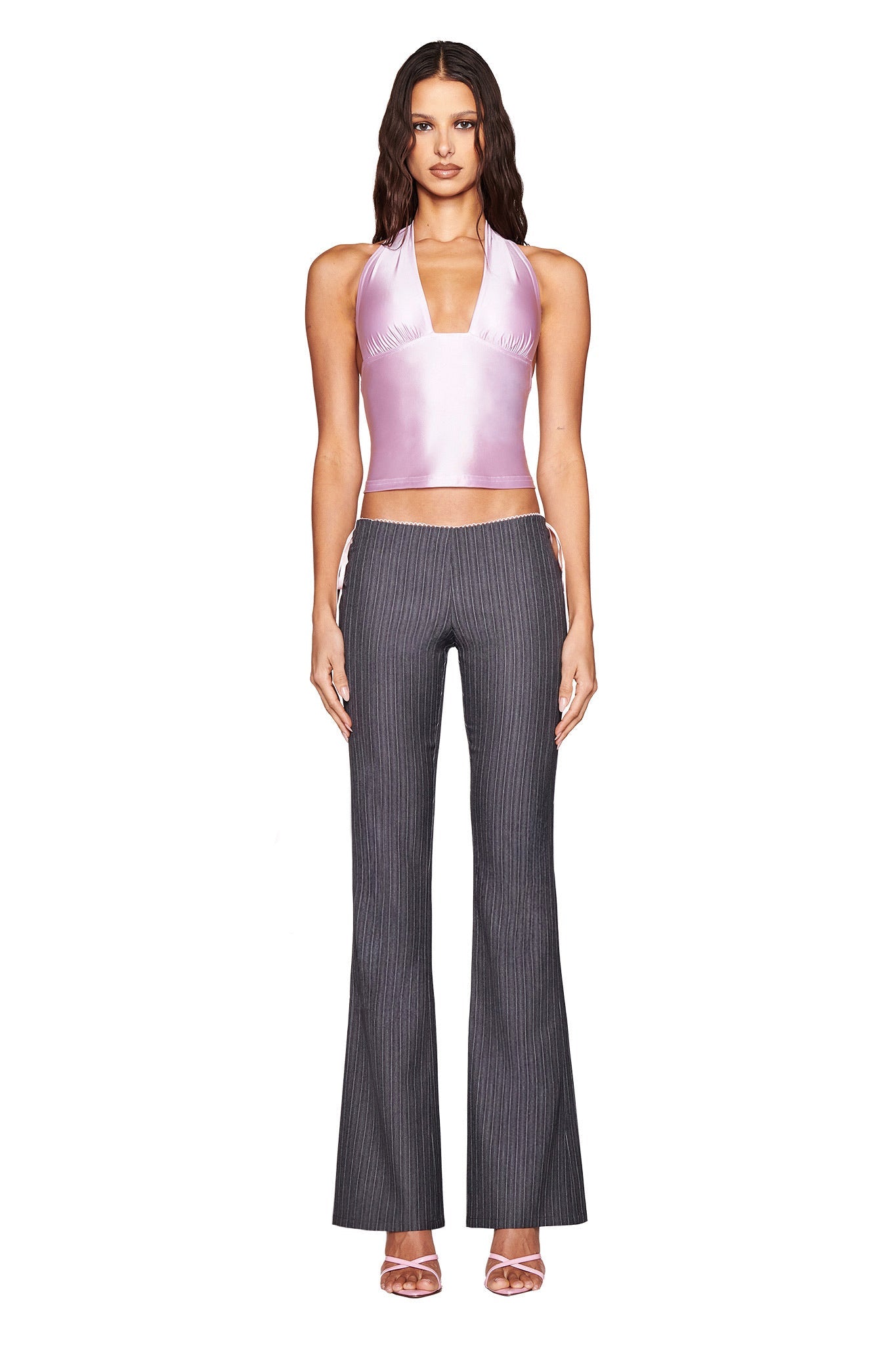 In a chic pose, they wear I.AM.GIA’s AURELIA HALTER TOP in baby pink with a bodycon fit, paired with dark pinstripe flared pants. Their long, wavy hair elegantly cascades against the plain white background, completing the stylish look.