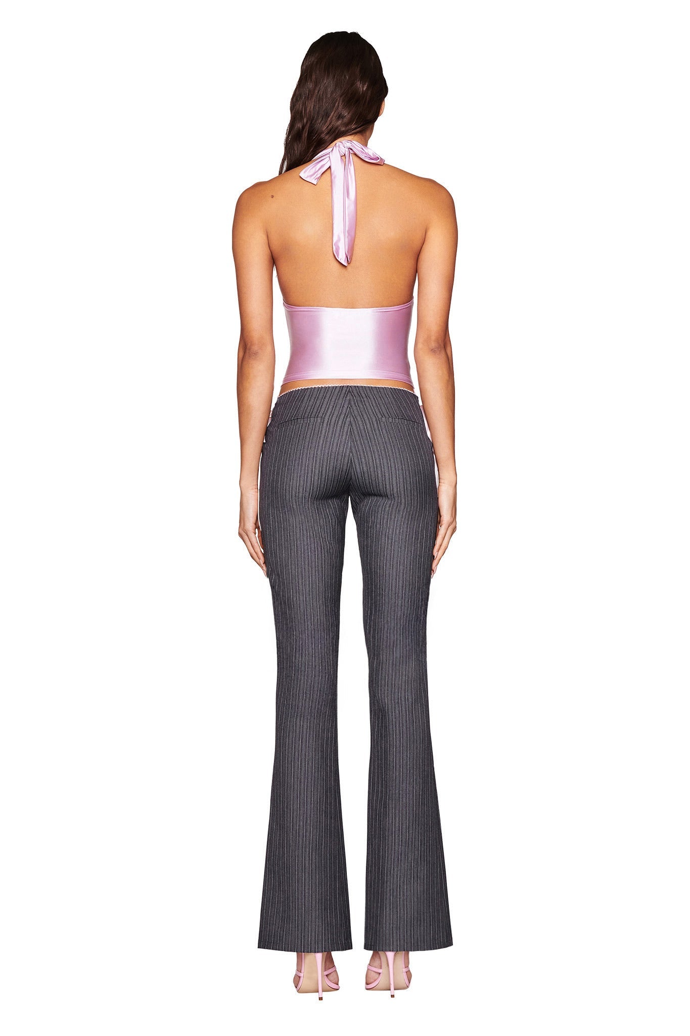 A person with long, wavy brown hair is wearing the AURELIA HALTER TOP - PINK by I.AM.GIA, featuring a ribbon tie, and high-waisted gray pinstriped pants. They are standing against a plain white backdrop.