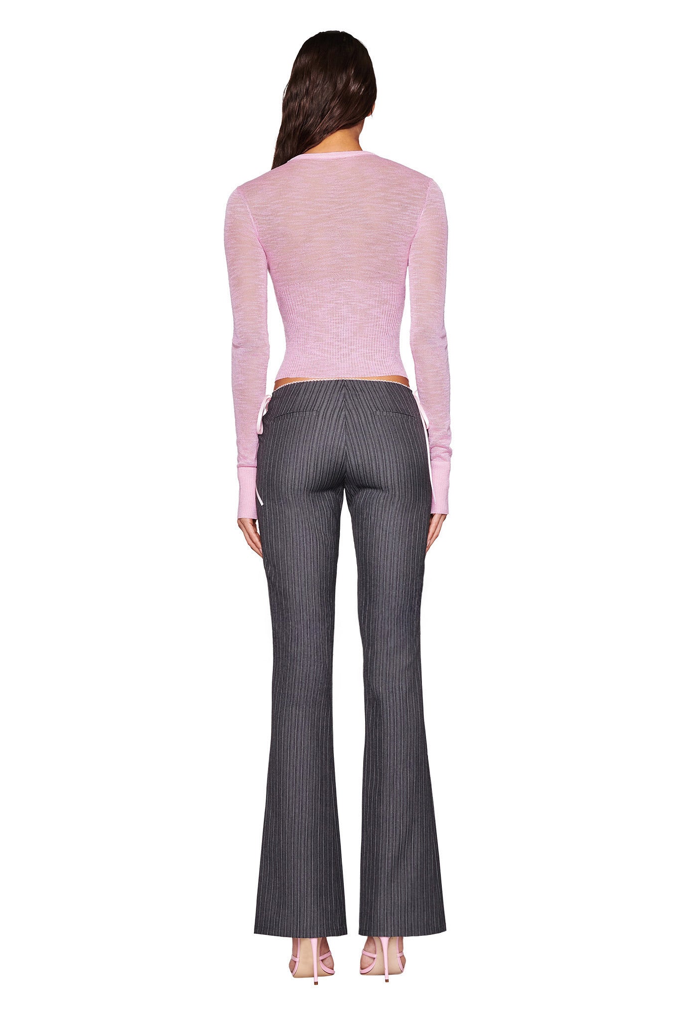 A person wears the I.AM.GIA MARIPOSA TOP in pink, featuring an open knit butterfly design, paired with gray pinstripe flared pants and matching pink high heels, set against a plain white background.