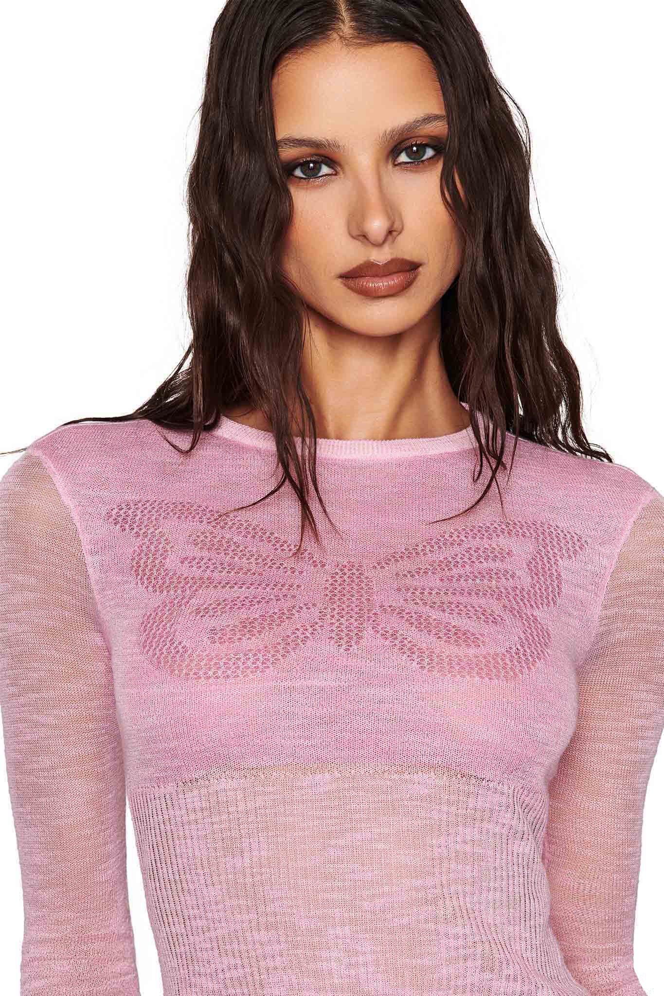 A woman with long, wavy hair wears the I.AM.GIA MARIPOSA TOP in pink, showcasing its open knit design and butterfly motif. She has a neutral expression and minimal makeup while standing against a plain white background.