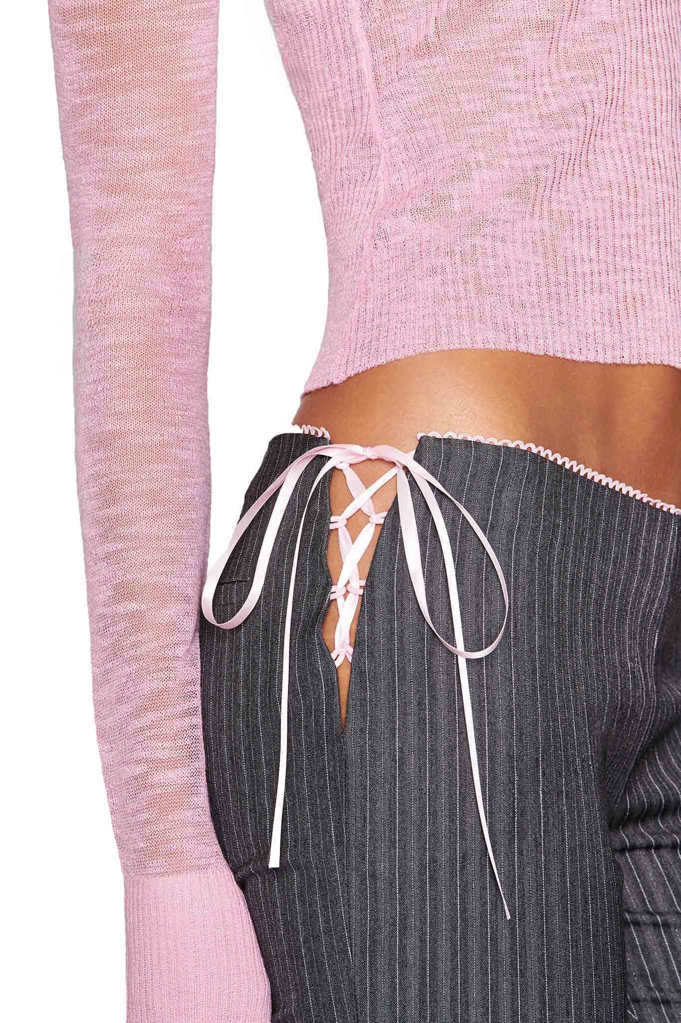 The image captures the midsection of a person wearing a pink long-sleeve top and the I.AM.GIA FLORIAN PANT - GREY pinstripe pants with flared legs, side lace-up detail, and pink ribbon, beautifully highlighting their texture and design.