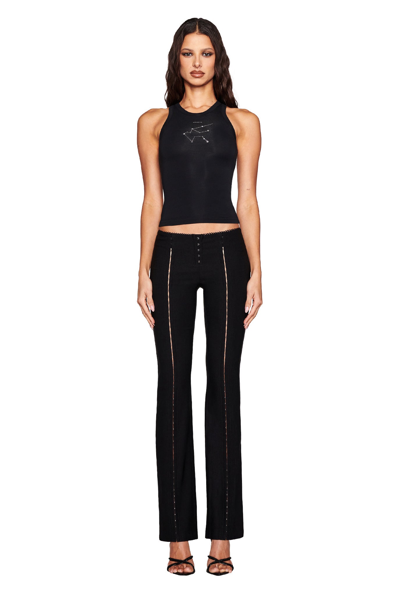 A person with long hair stands facing forward in a sleeveless black top and I.AM.GIA's RONAN PANT - BLACK, crafted from stretch woven material with stitching details. They're also wearing black open-toed shoes. The plain white background highlights their sleek ensemble.