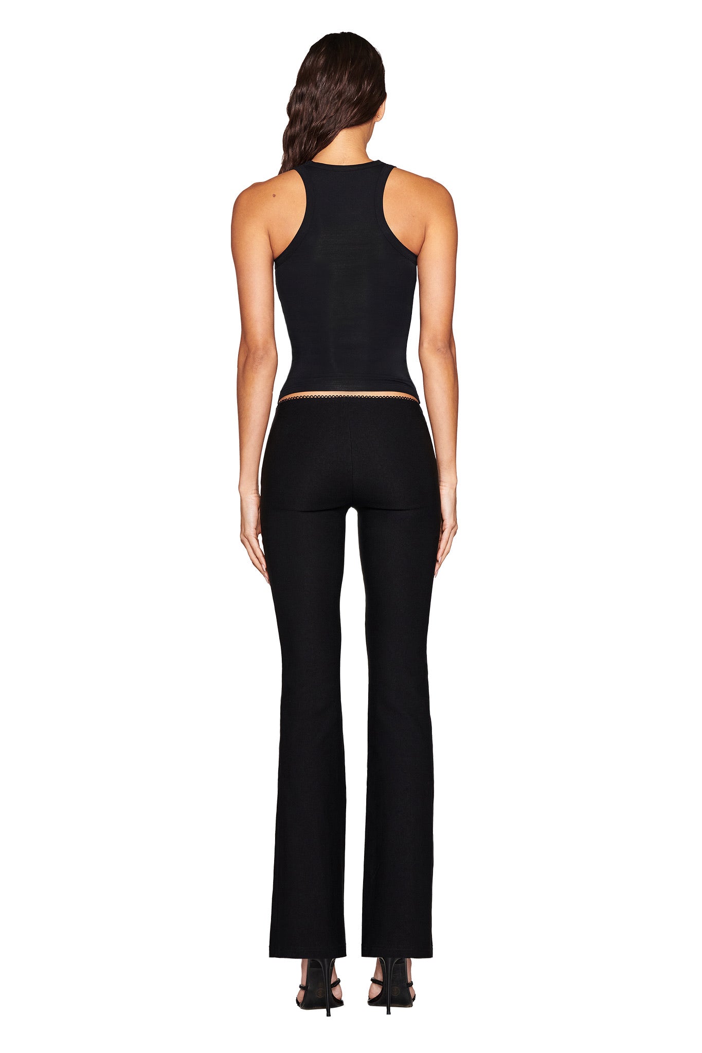 A woman with long brown hair is seen from behind wearing the I.AM.GIA fitted black sleeveless top and RONAN PANT - BLACK, made of stretch woven material. She's posing against a white background in high-heeled shoes, highlighting the sleek lines of her ensemble.