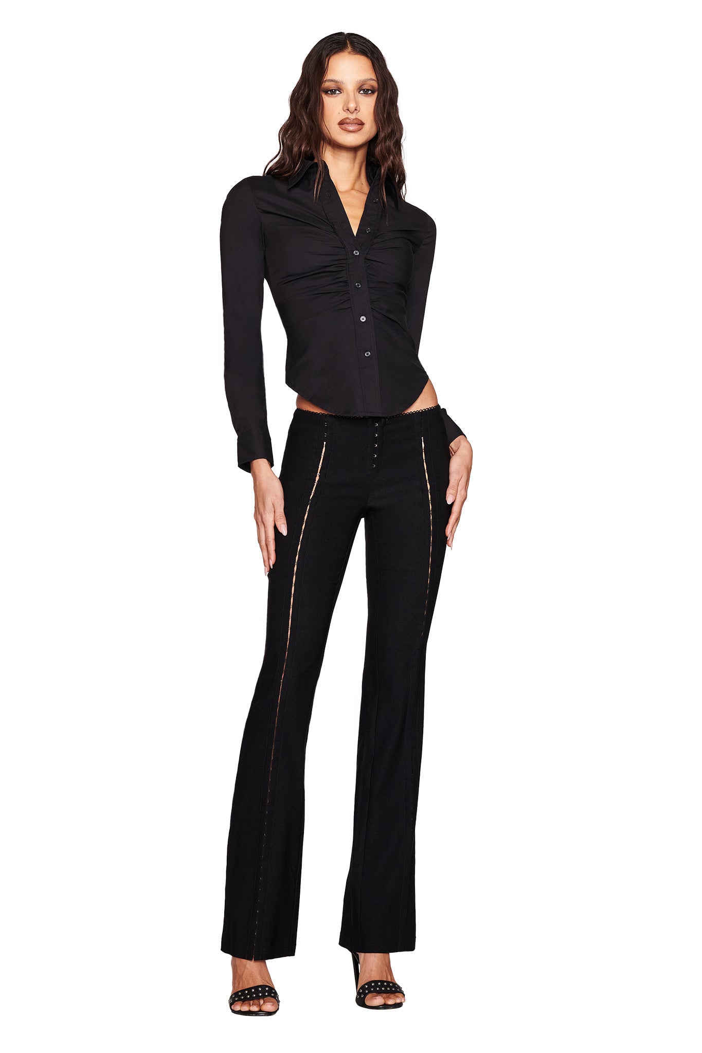 A person confidently models the RONAN PANT - BLACK by I.AM.GIA, featuring high-waisted design and decorative seams. The fitted black long-sleeve blouse complements the stretch woven fabric, and strappy black heels complete the stylish look against a plain white backdrop.