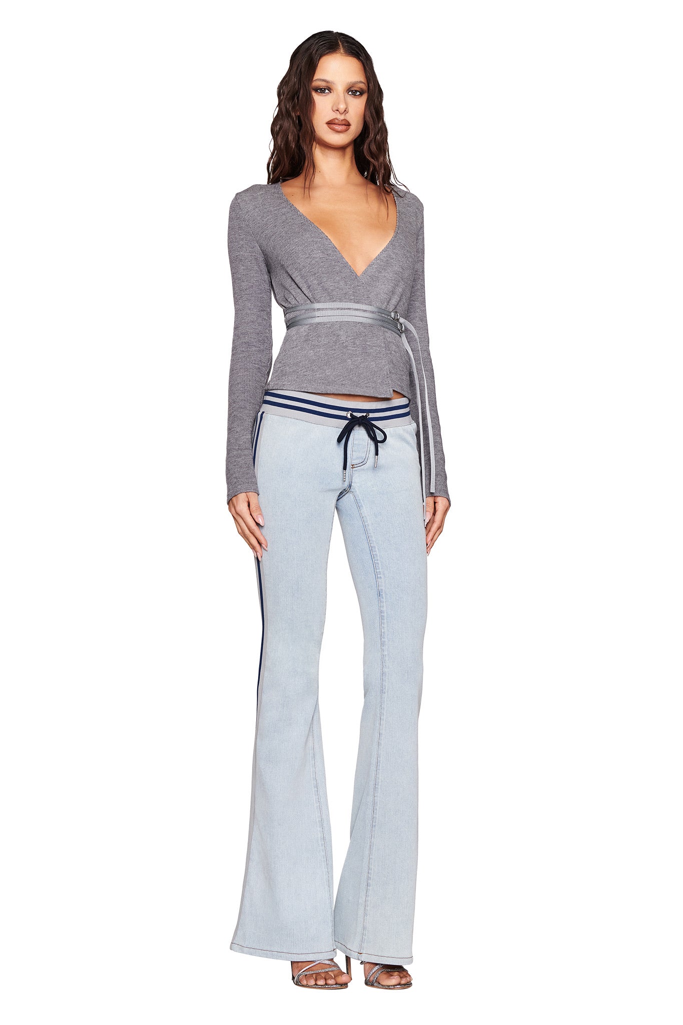 A person stands in a deep V-neck gray wrap-around top with a waist tie, paired with I.AM.GIA's LENNOX JEAN - BLUE featuring side stripes and light wash denim. Strappy heels complete the look against a plain white background.