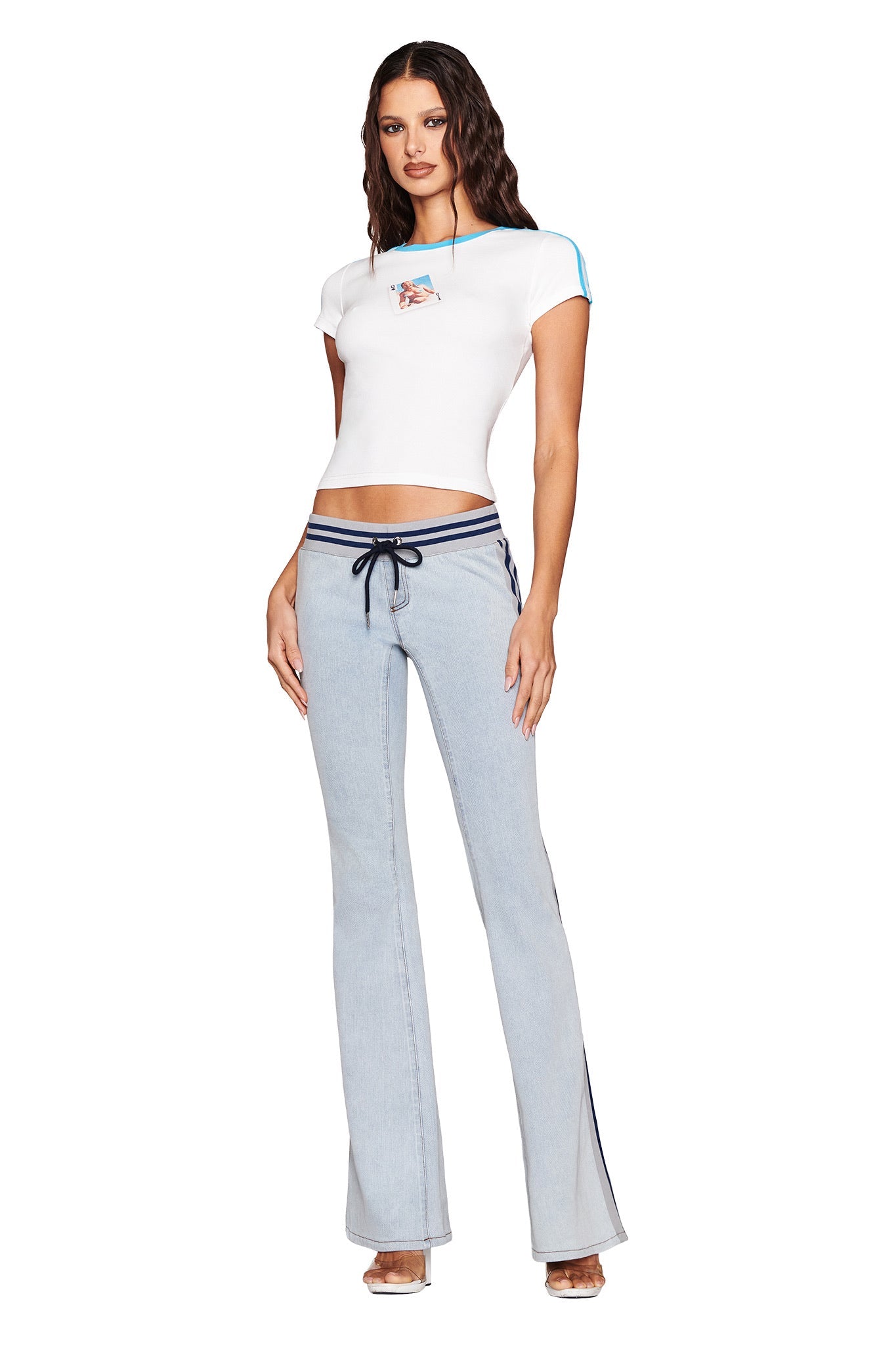 A woman poses in an I.AM.GIA white crop top, LENNOX JEAN in denim blue, and clear high heels. She stands against a white background, her wavy hair cascading over her shoulders.