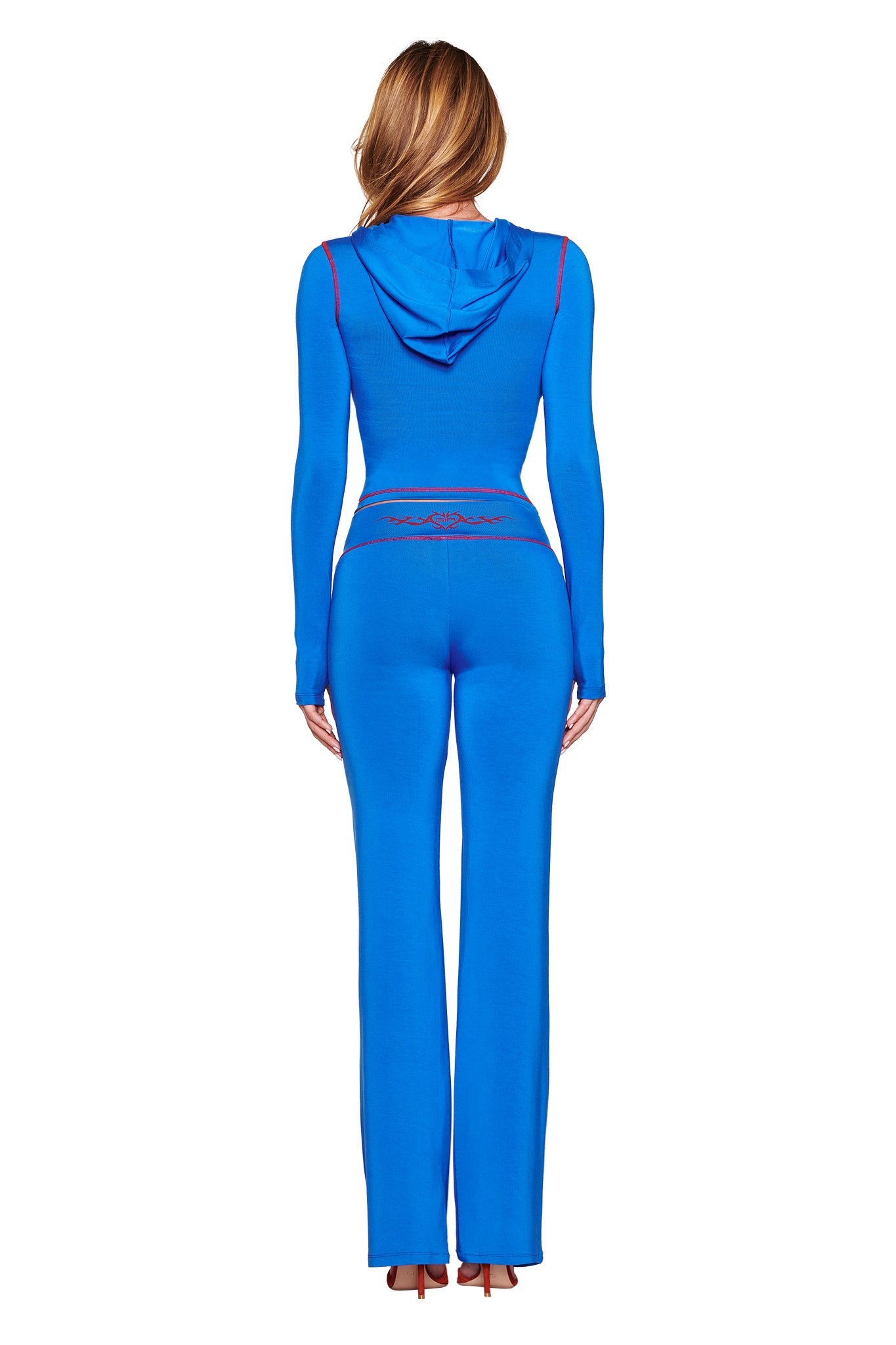 The long-haired woman is dressed in I.AM.GIA's Blare Trackpant 2.0 in royal blue with red accents, featuring a hood, paired with brown heeled shoes. This creates an elegant sporty-chic look from the back view.
