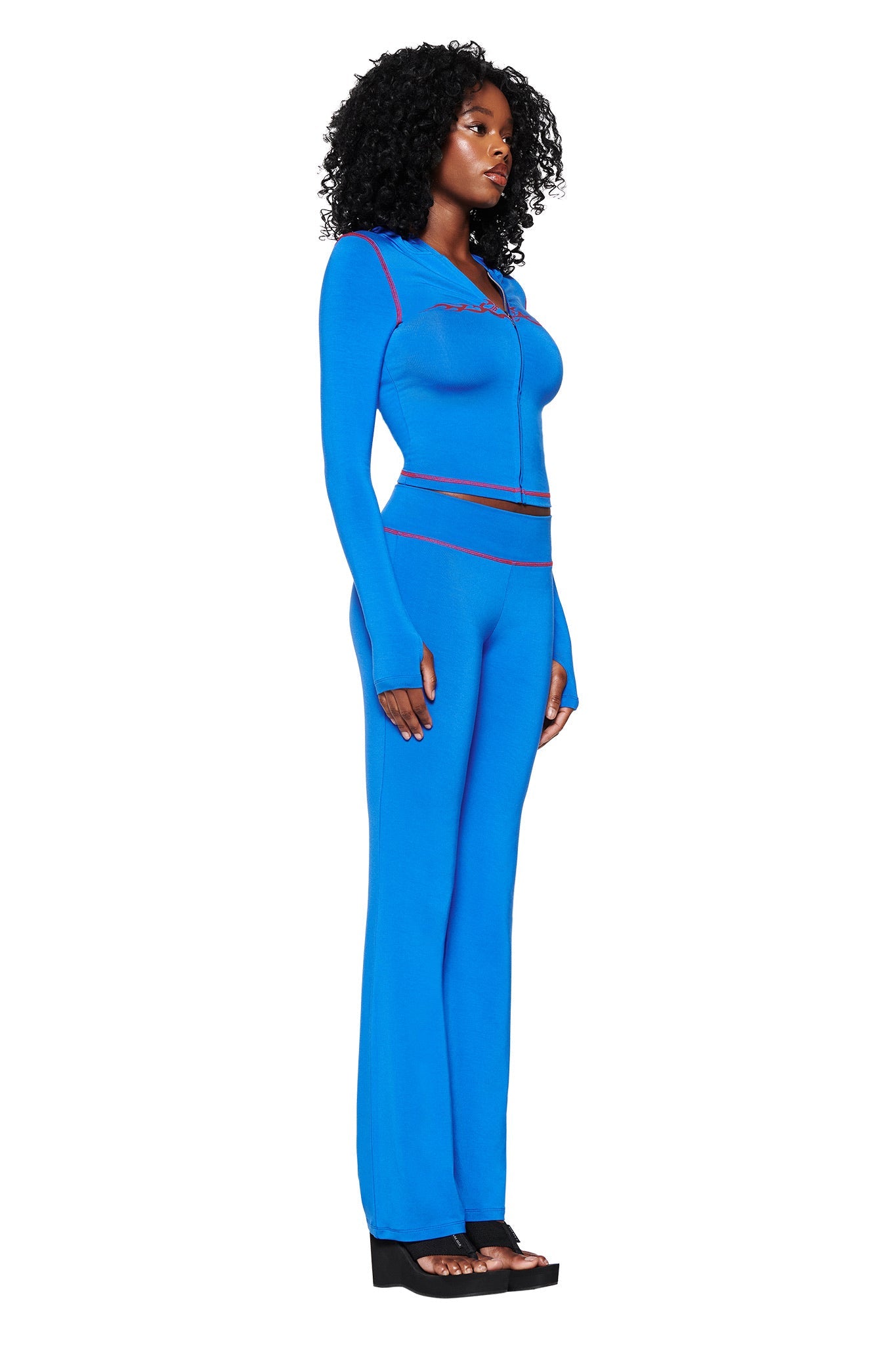 A woman in profile sports a royal blue long-sleeved top and I.AM.GIA's Blare Trackpant 2.0 in Royal Blue, both with red stitching. Her curly hair complements the style, paired with black platform sandals against a plain white backdrop.