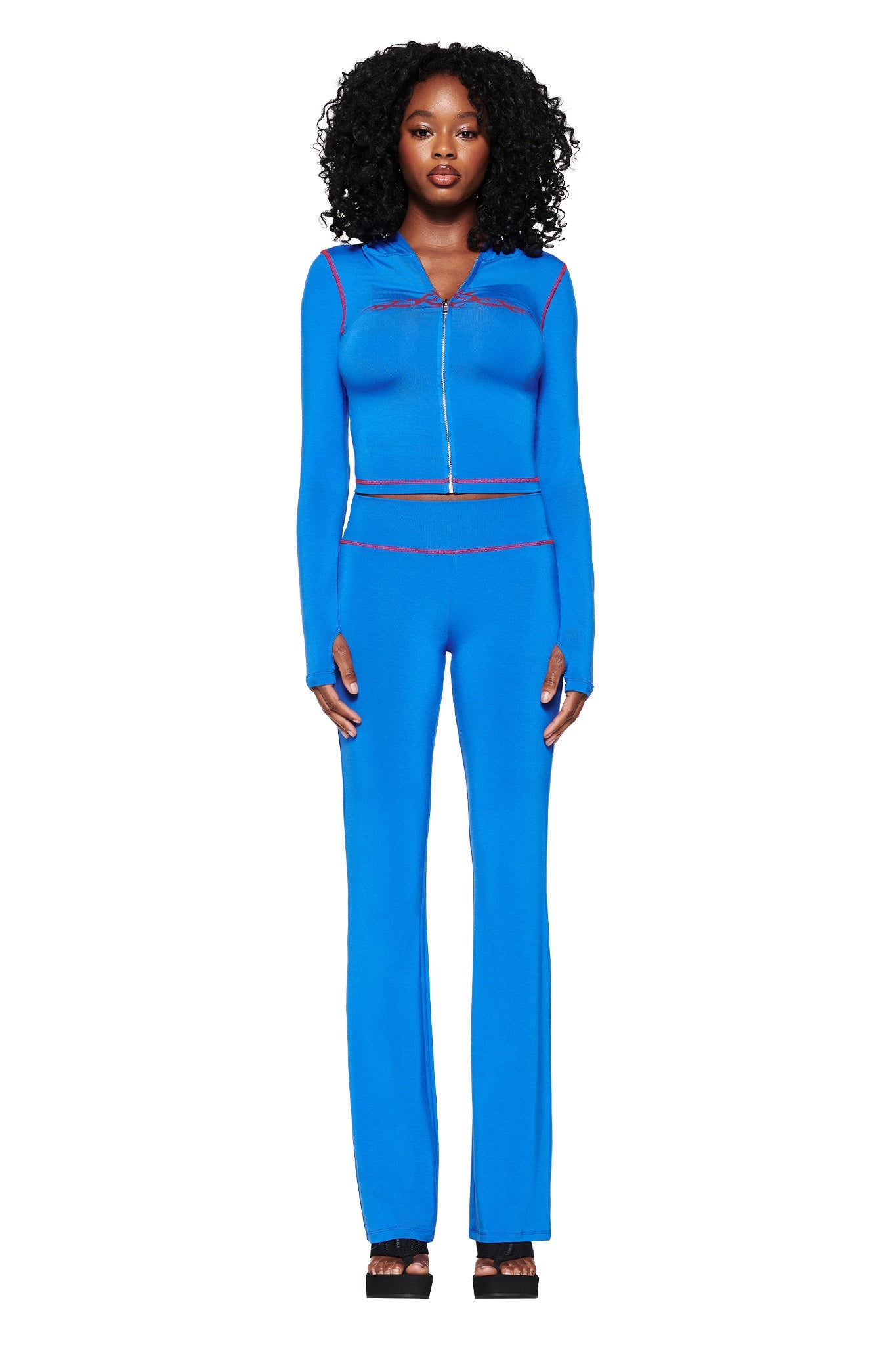 A person with curly hair faces forward wearing I.AM.GIA's Blare Trackpant 2.0 in royal blue, complemented by black platform shoes. Thin pink lines accentuate the bodycon silhouette against a plain white backdrop, highlighting their vibrant presence.