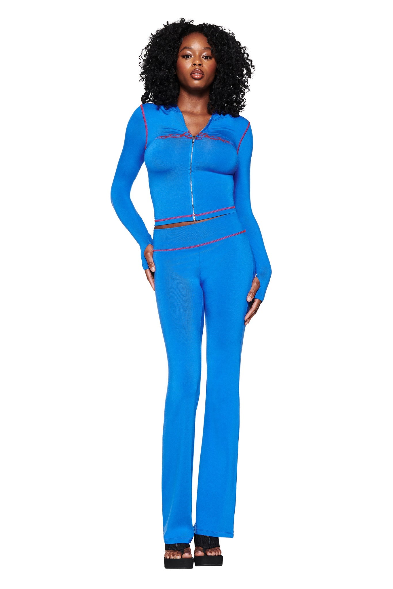 A woman with curly hair confidently poses in an I.AM.GIA Royal Blue form-fitting BLARE HOODIE featuring red trim and a front zipper. She stands against a white background, hands on hips, wearing black platform sandals.