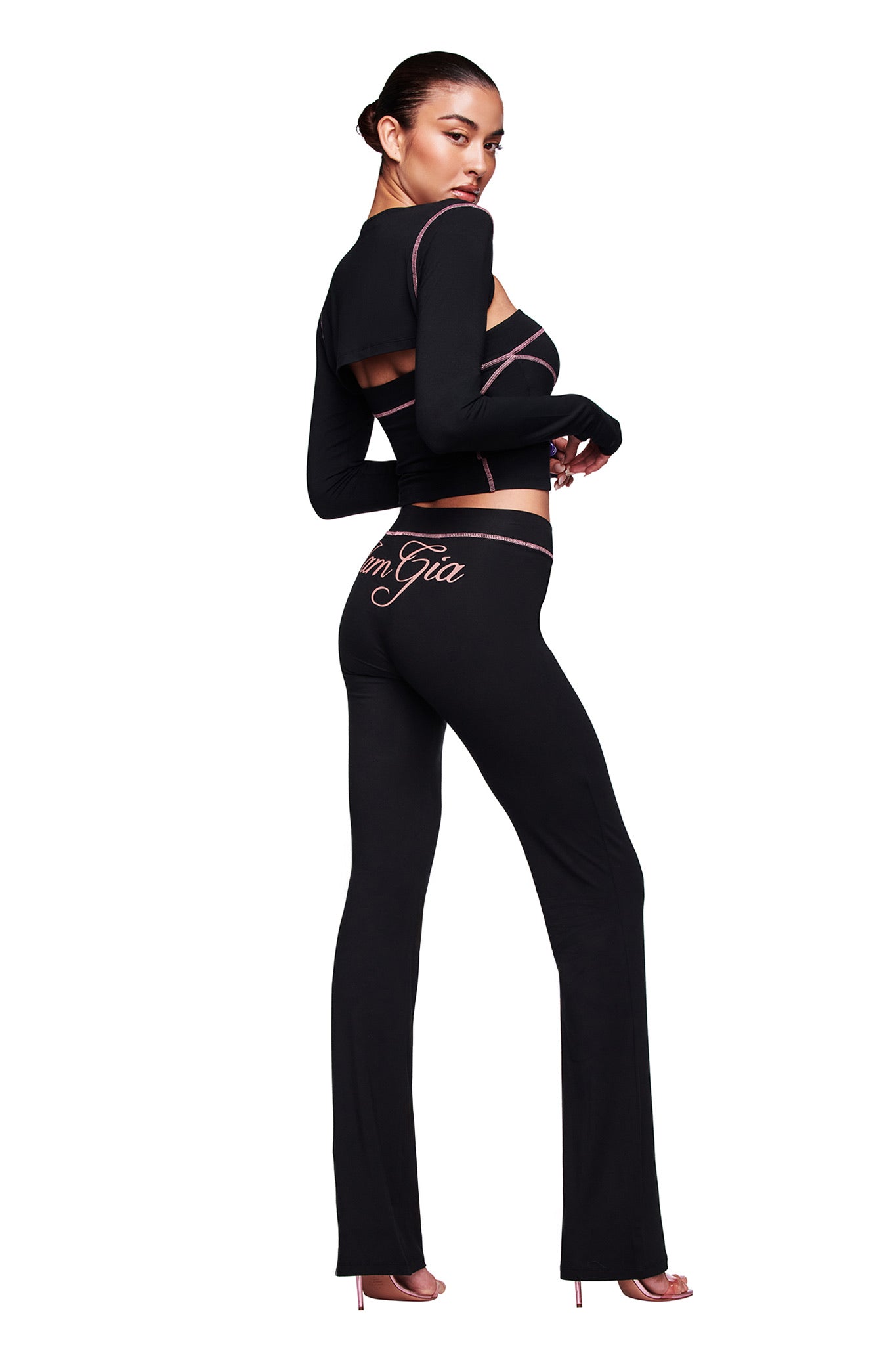A woman models the I.AM.GIA BLARE BOOB TUBE in black, paired with matching pants showing the brand logo on the back. She finishes the look with pink heels, all set against a white backdrop.
