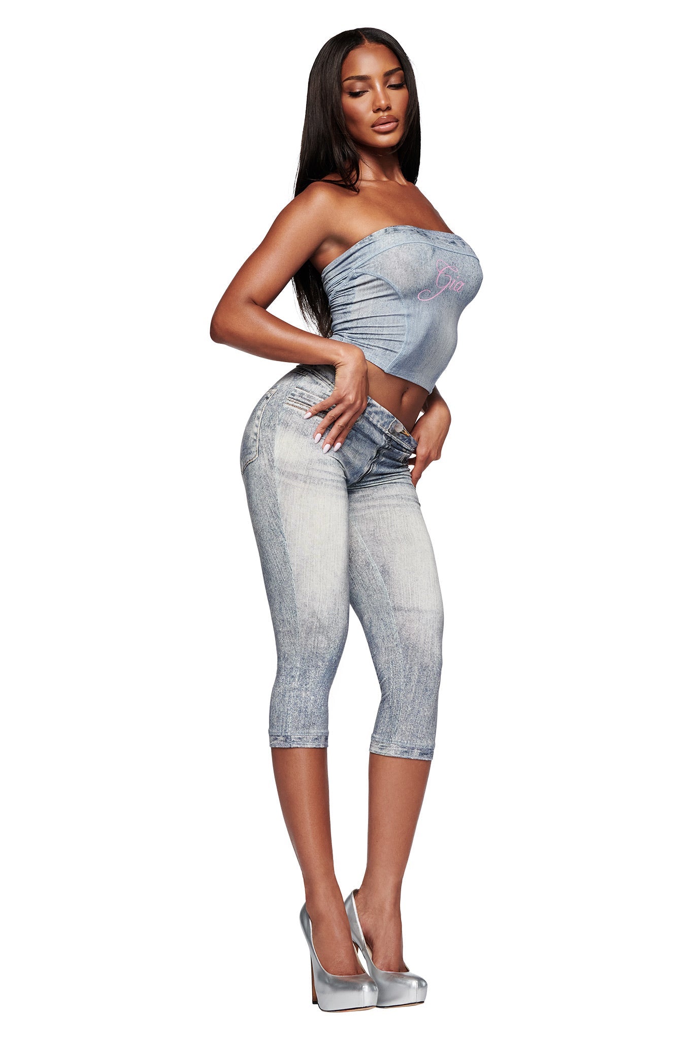 A woman wears the I.AM.GIA BLARE BOOB TUBE - DENIM TROMPE L'OEIL crop top with matching high-waisted capri pants and silver heels, posing against a white background.