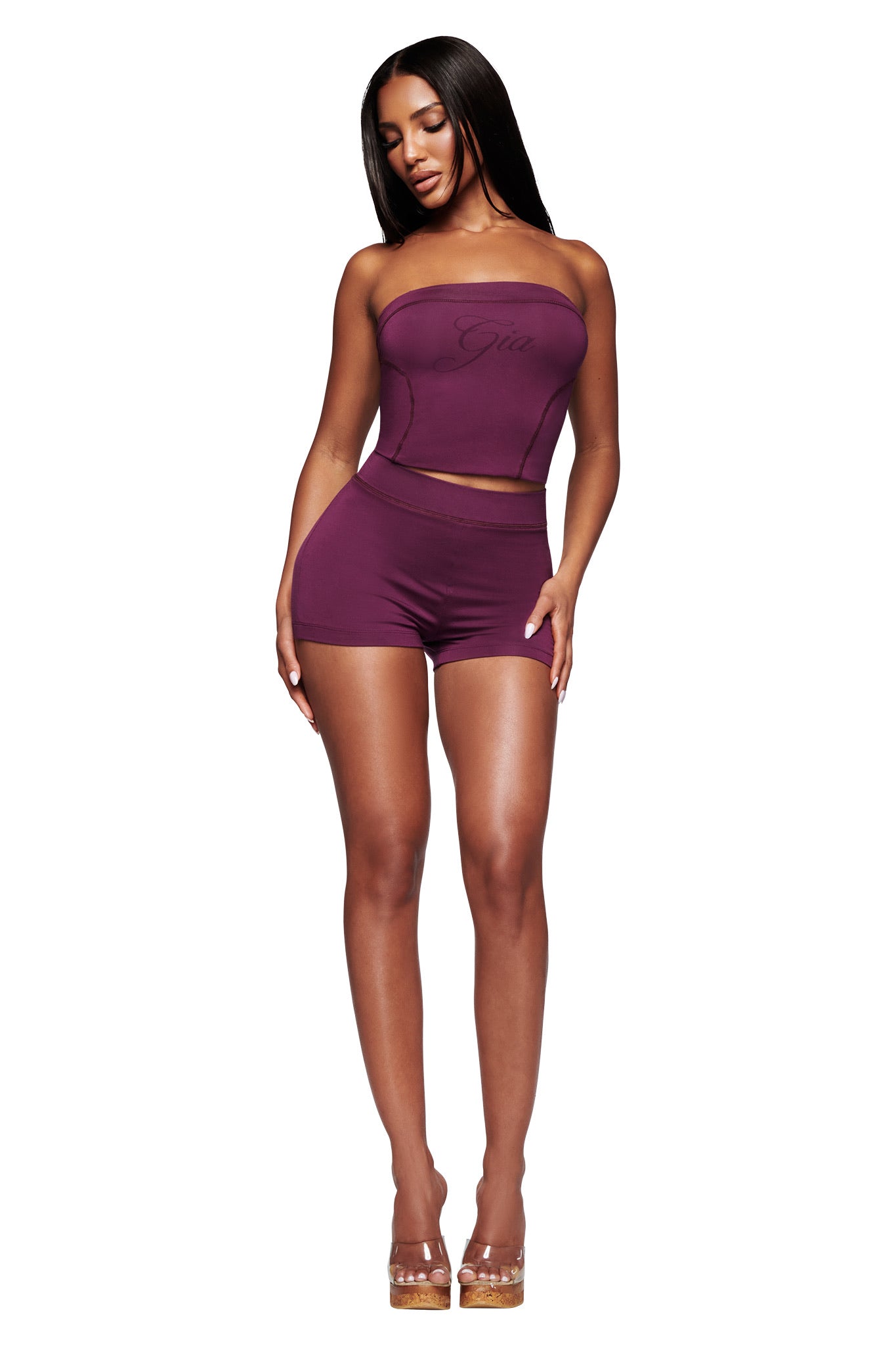 A woman wears the I.AM.GIA BLARE BOOB TUBE in plum with matching shorts and clear high-heeled shoes, making a bold, modern statement against a white background.