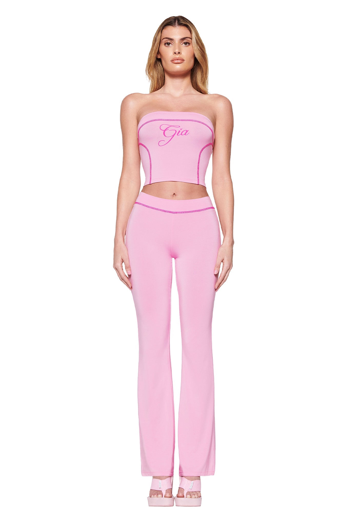 A person stands against a white background in bubblegum pink hues, wearing a strapless top with the I.AM.GIA logo and matching BLARE TRACKPANT - PINK: BUBBLEGUM, complemented by platform heels, radiating chic confidence.