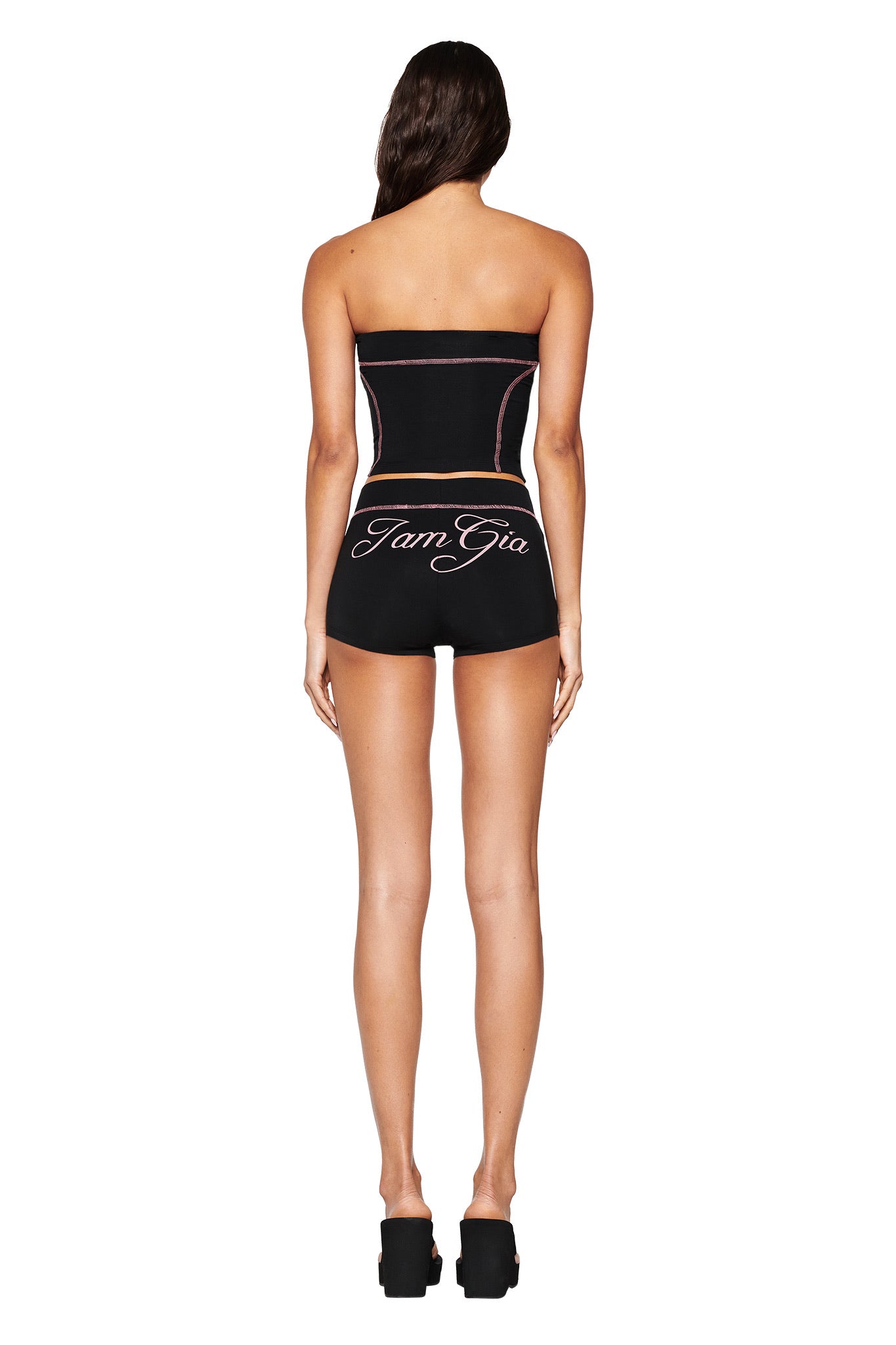 From the back, a person with long brown hair wears I.AM.GIA's BLARE BOOB TUBE in black and matching shorts with logos, set against a white backdrop. Black platform shoes enhance the sporty seams of their outfit.