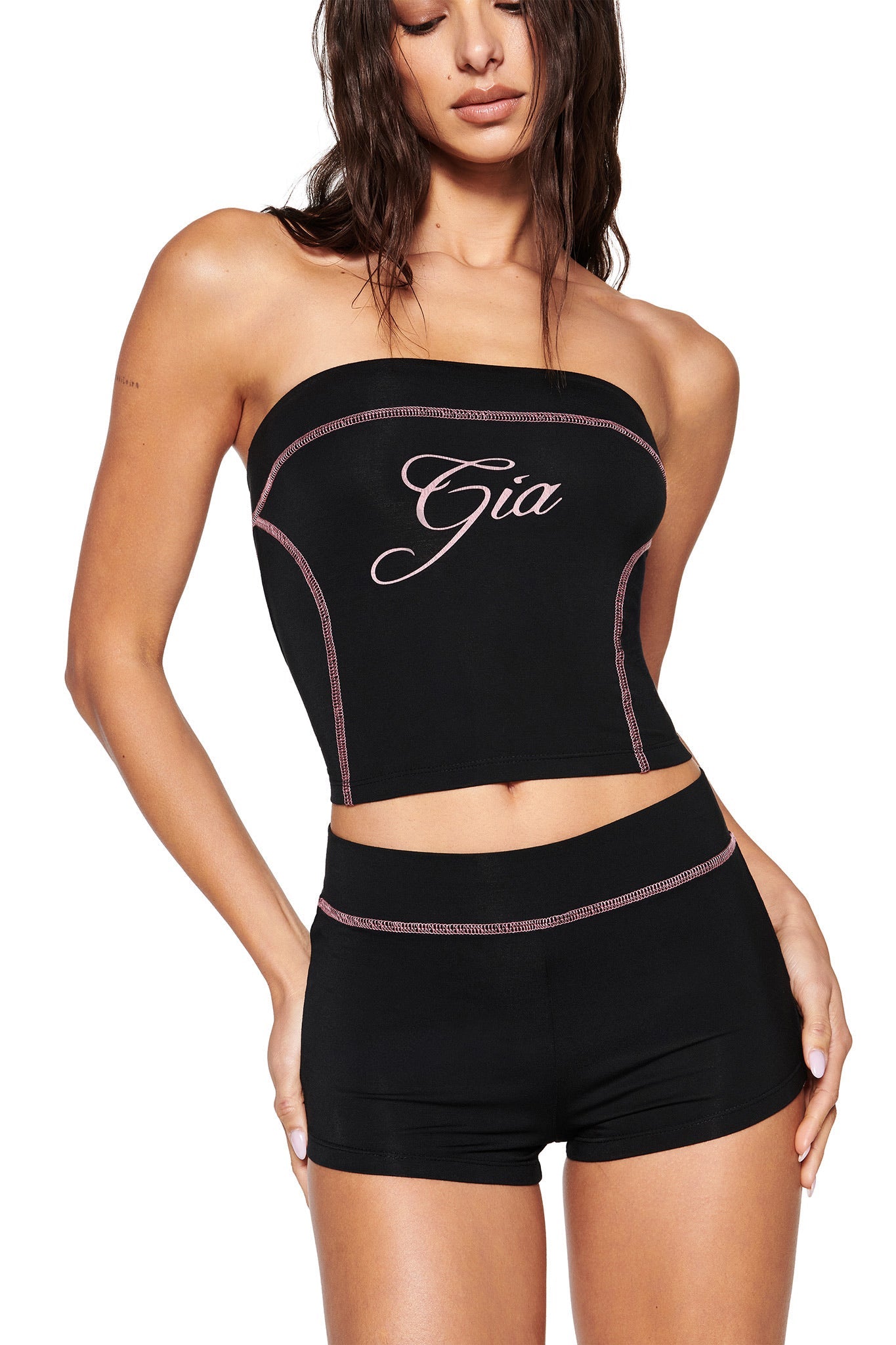 Wearing I.AM.GIA's BLARE BOOB TUBE - BLACK adorned with pink "Gia" script and matching sporty seam shorts, a person poses confidently with long dark hair cascading, hand on hip, embodying the brand's edgy style.