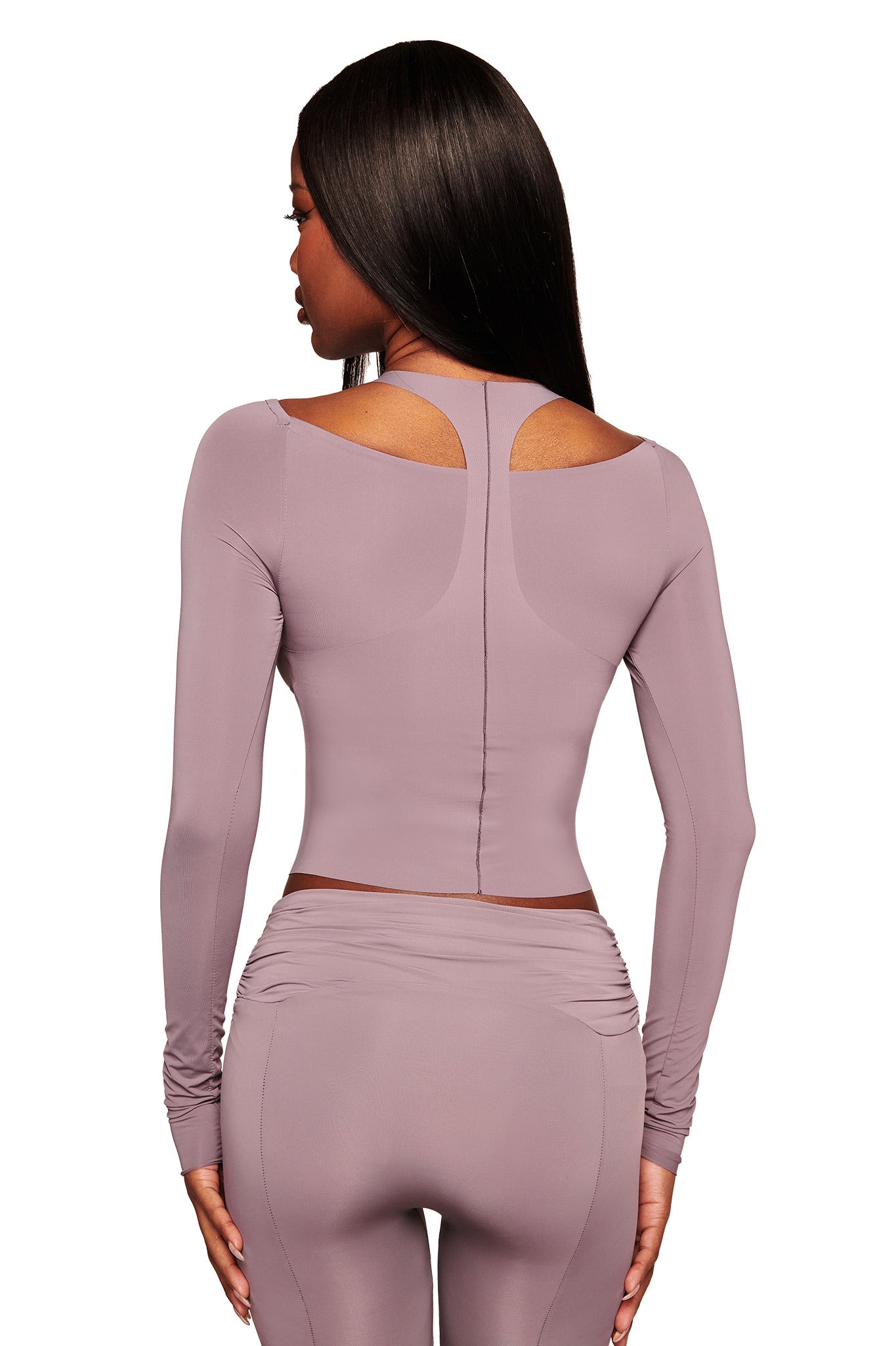 A woman with straight dark hair wears the I.AM.GIA NERINA LONG SLEEVE TOP in dusty lilac, featuring shoulder cutouts and a racerback, paired with matching high-waisted leggings. She is seen from behind, gazing slightly to the side.
