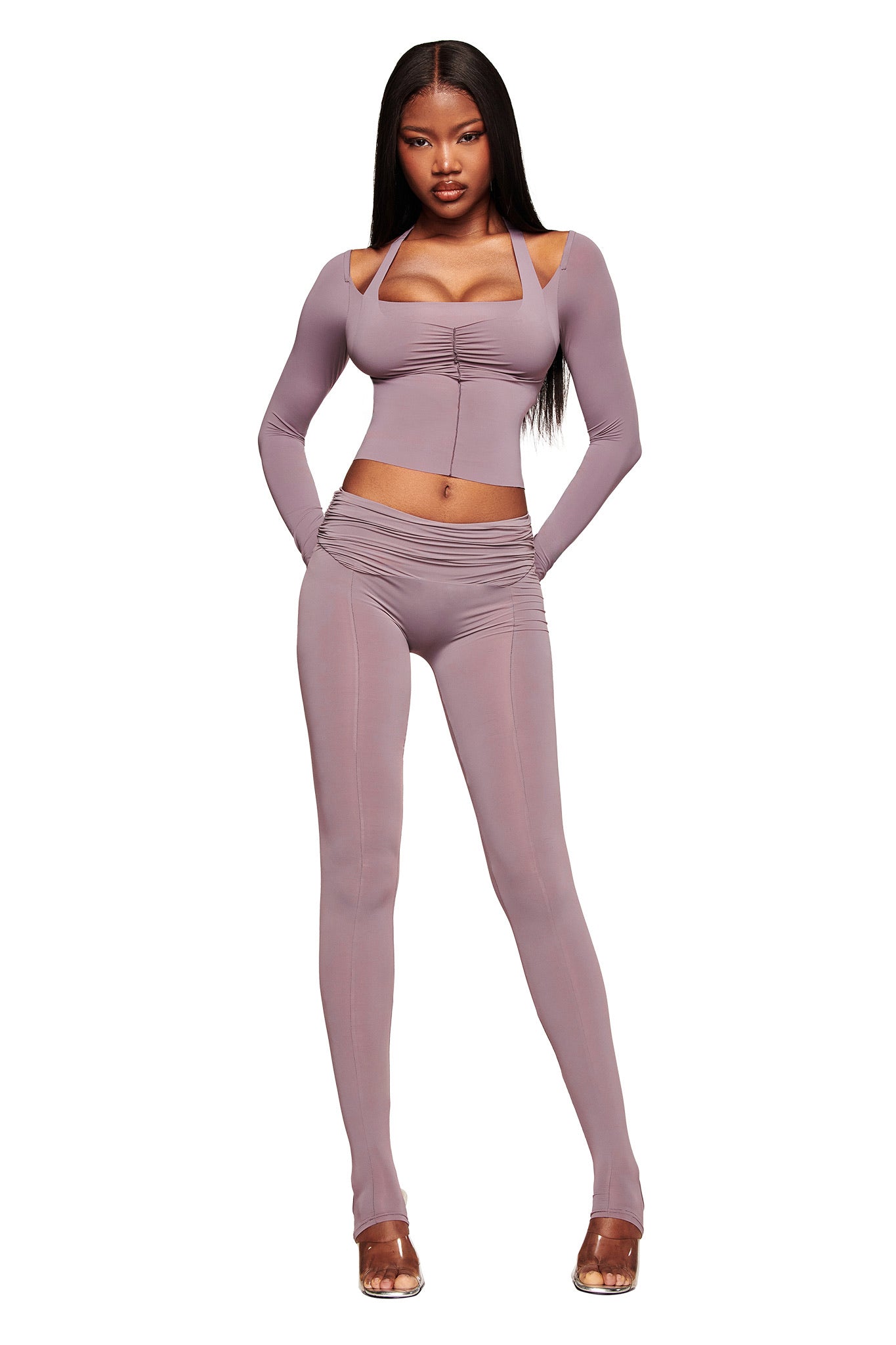 A woman models the I.AM.GIA NERINA LEGGING in dusty lilac, a two-piece outfit with long sleeves, full-length leggings, cut-out shoulders, and ruched details. She stands against a white background in clear heels.