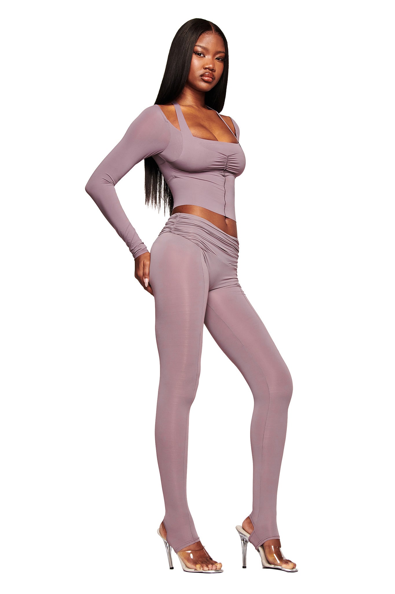 A woman wears I.AM.GIA's NERINA LEGGING in dusty lilac with a matching fitted long-sleeve crop top and clear high-heeled sandals, posing against a white background.