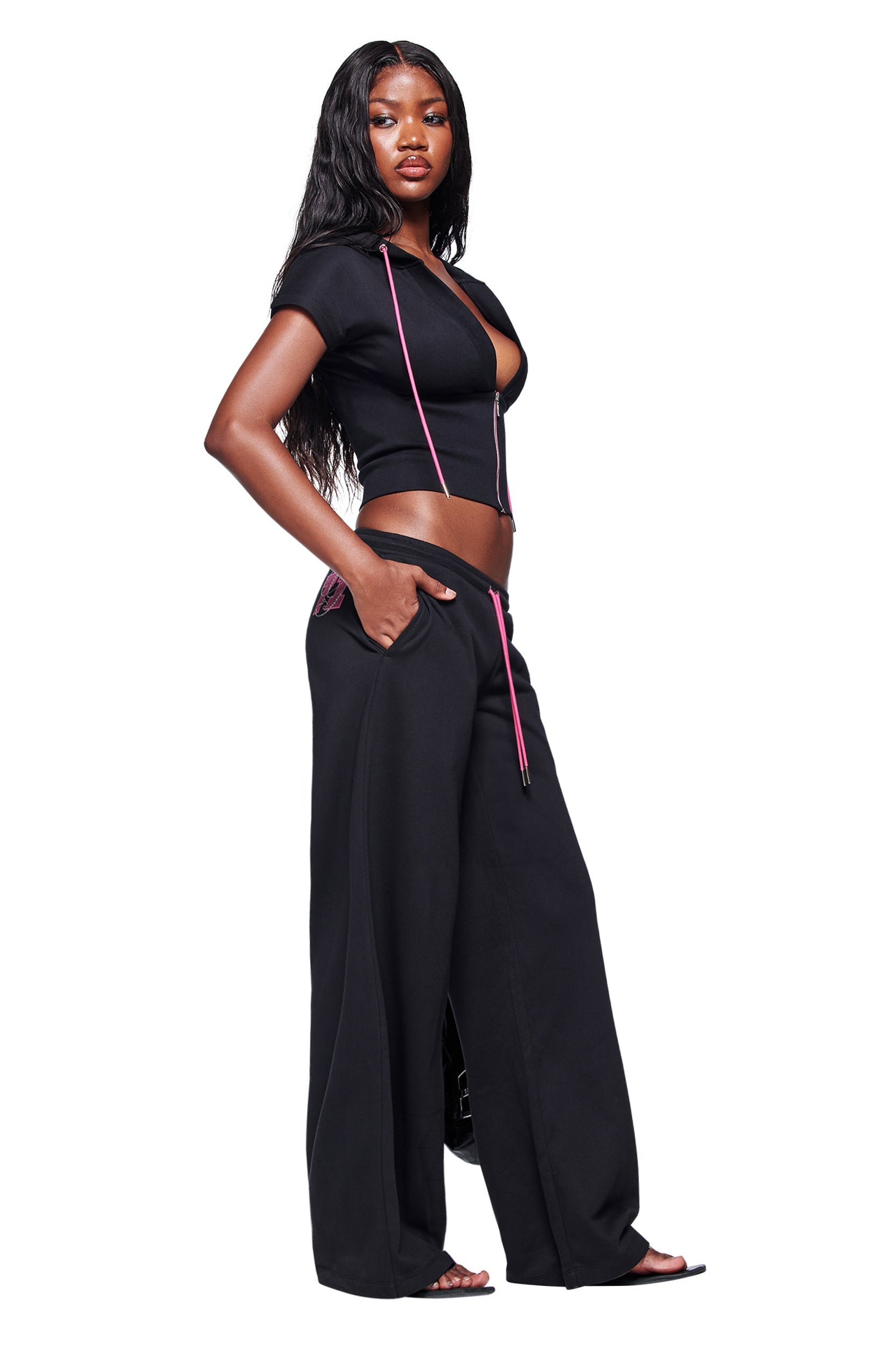 A woman with long wavy hair wears a short-sleeved black crop top with pink drawstrings and the I.AM.GIA ARLO TRACK PANT - BLACK, standing in a relaxed pose with her hands in her pockets.