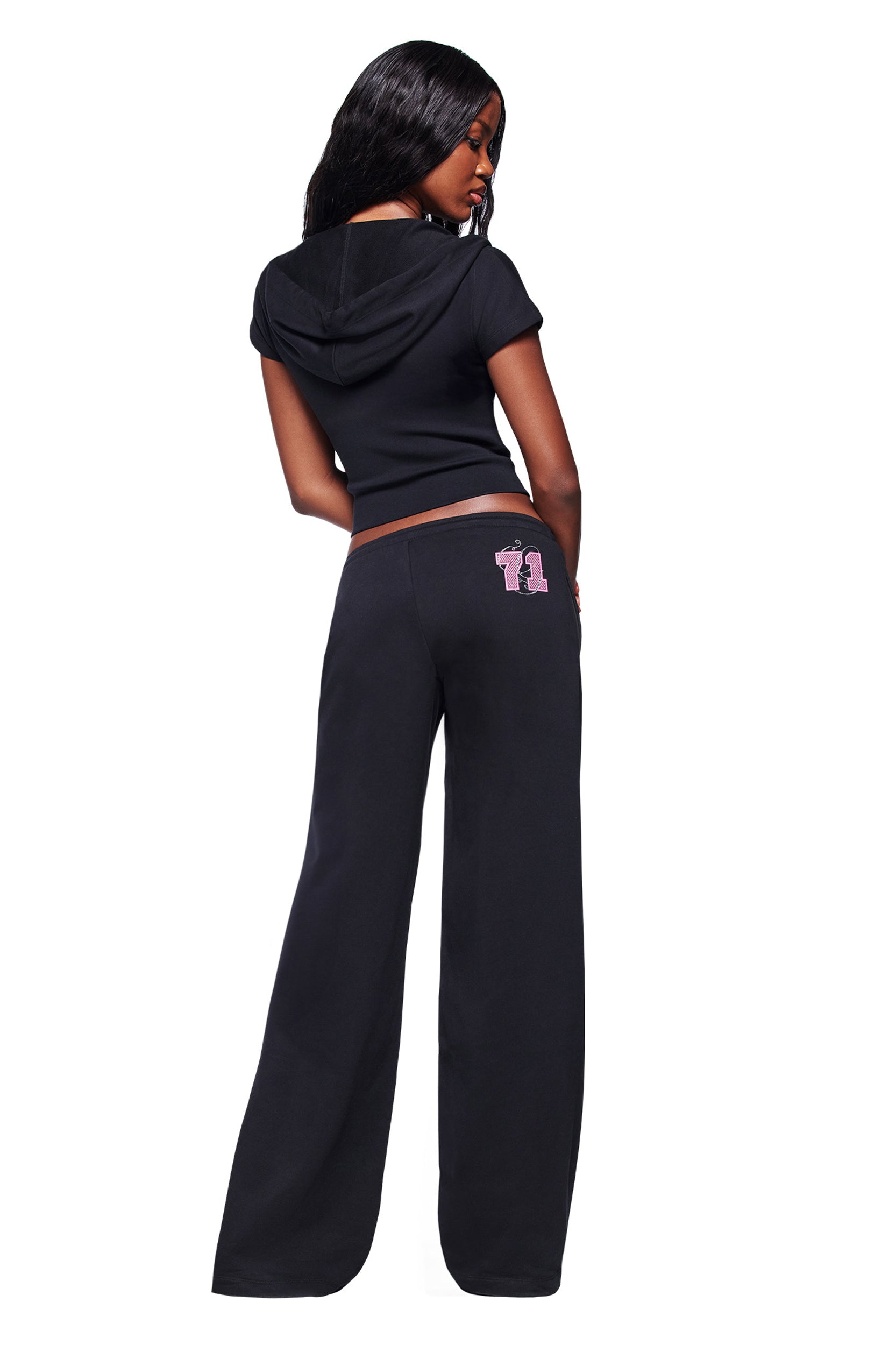 A woman with long, dark hair models the I.AM.GIA ARLO TRACK PANT in black, featuring a pink graphic on the back pocket. She faces away from the camera against a white background, wearing matching black separates.