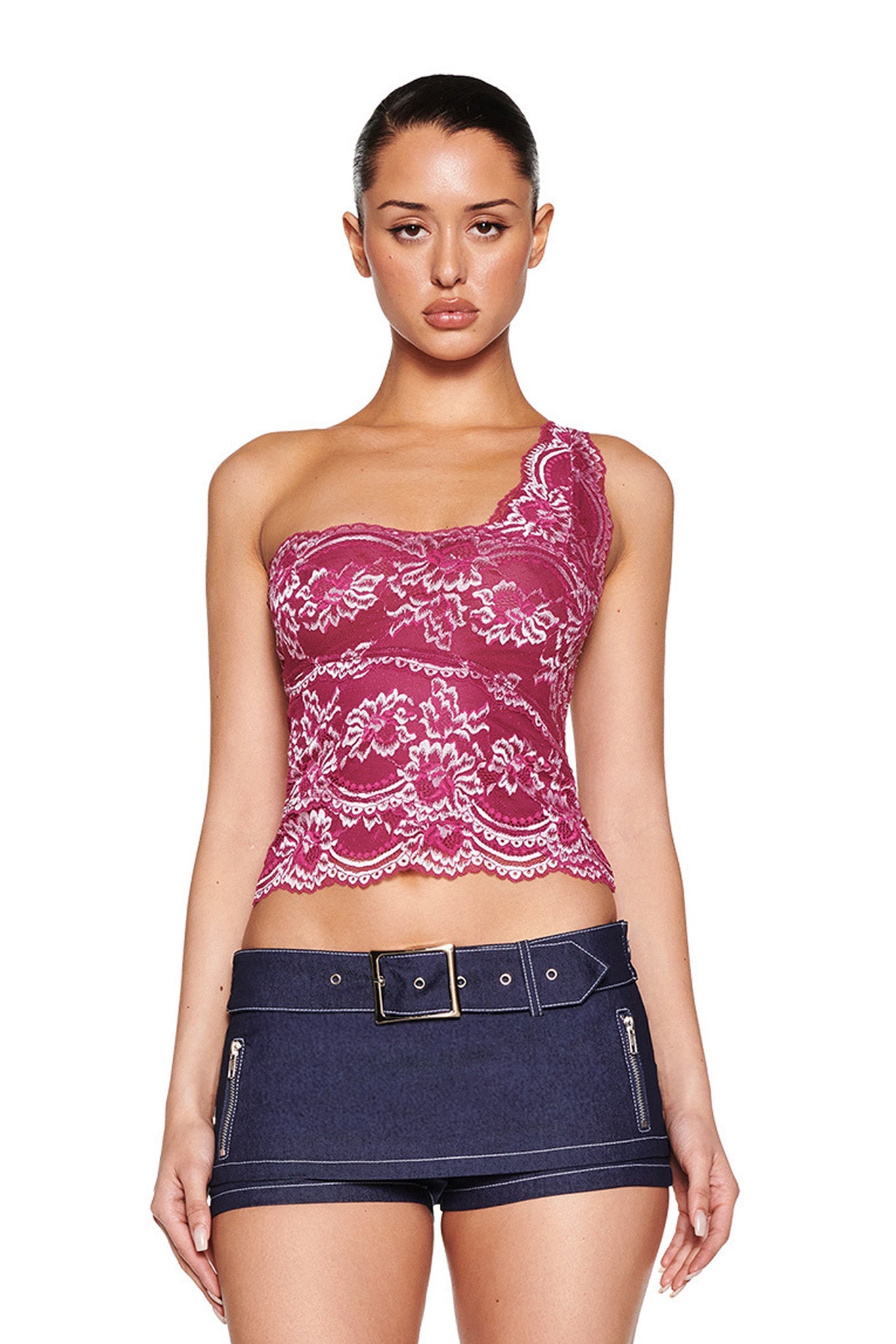A person poses against a white background, wearing I.AM.GIA's PHOENIX TOP in Purple Plum and a dark denim mini skirt featuring zipper details and a belt. Their hair is tied back as they look forward.