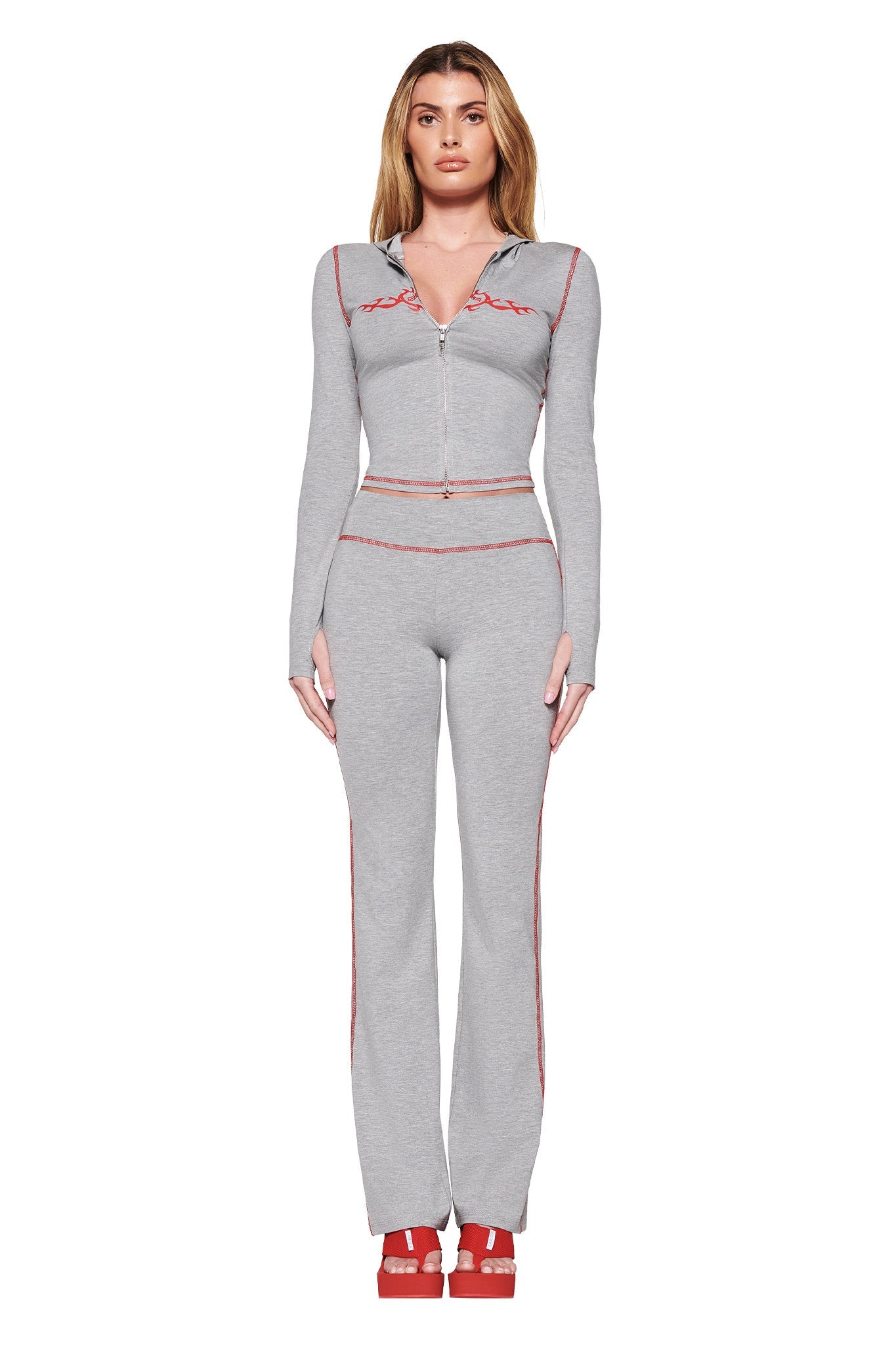 A person is wearing a fitted tracksuit—dove grey Blare Trackpant 2.0 by I.AM.GIA, featuring red stitching and the GIA Flame logo. The outfit includes a long-sleeve zip-up top with matching pants, paired with red platform shoes against a white background.