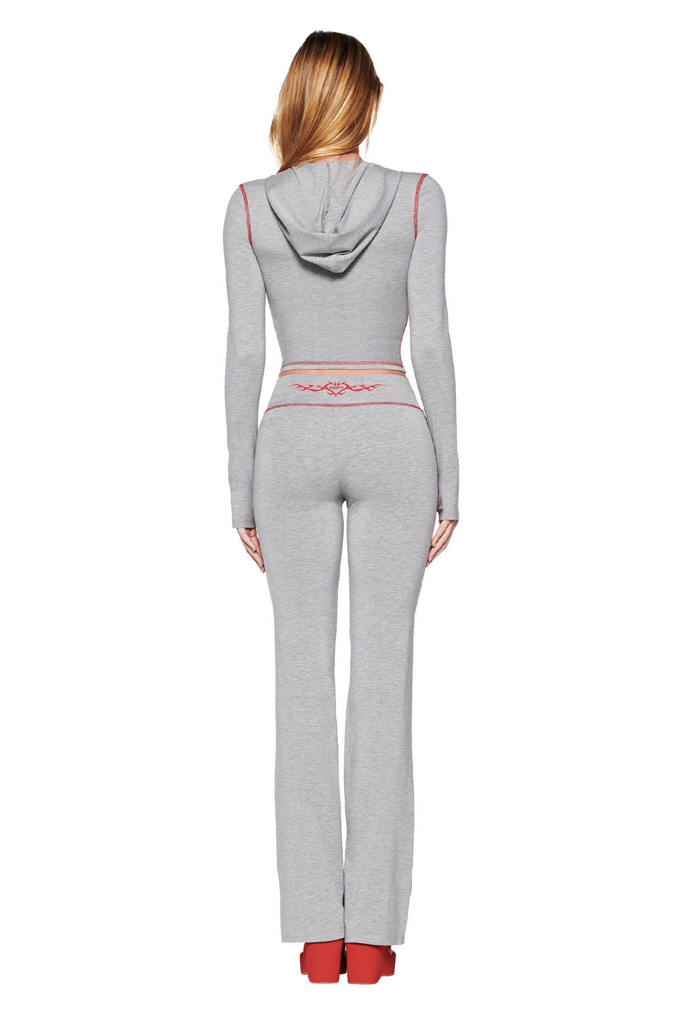 The rear view shows a person with long hair in a dove grey, form-fitting hoodie and BLARE TRACKPANT 2.0 - GREY: DOVE GREY, featuring red accents. Red shoes complete the look. The GIA Flame logo is visible on the back of the hoodie and pants waistband from I.AM.GIA.
