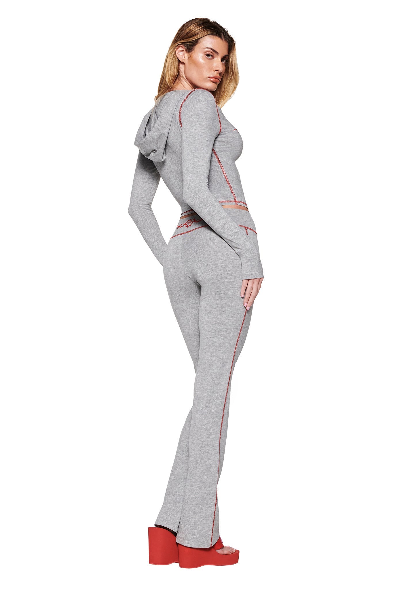 A person in a fitted I.AM.GIA Blare Trackpant 2.0, dove grey with red accents and the GIA Flame logo, stands sideways. The outfit features a hooded top and flared legs paired with high platform red sandals against a crisp white background.