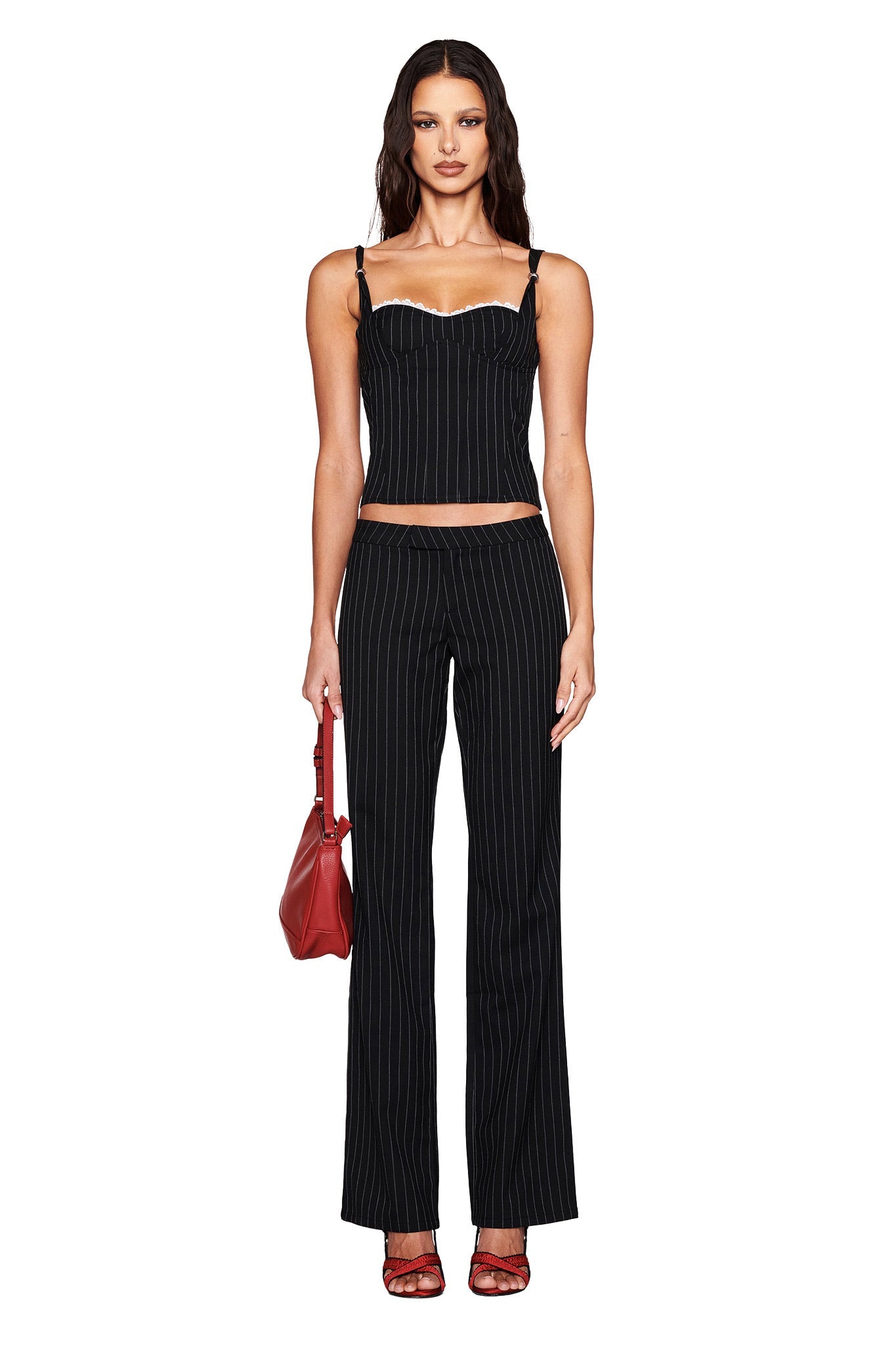 A person stands against a white backdrop, showcasing an under-bust look with a fitted I.AM.GIA MARRION Corset in black and matching flared pants. They hold a red handbag and wear matching red shoes, their long wavy hair completing the chic ensemble.