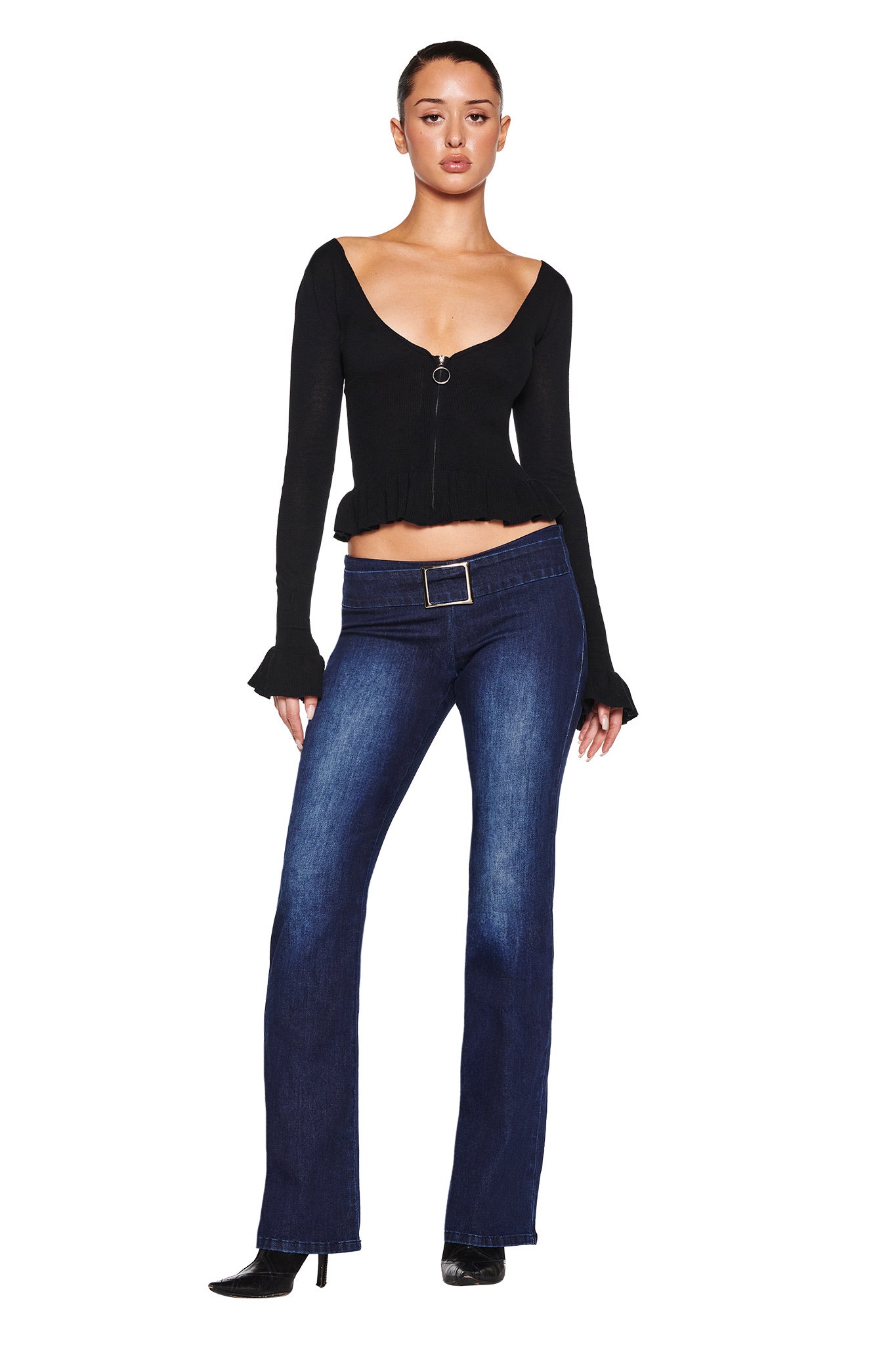 Someone wearing the I.AM.GIA RAVEN TOP in black with a deep V neckline pairs it with dark blue bootcut jeans. They stand against a white background, complete the look with black pointed-toe shoes, and have their hair neatly tied back.