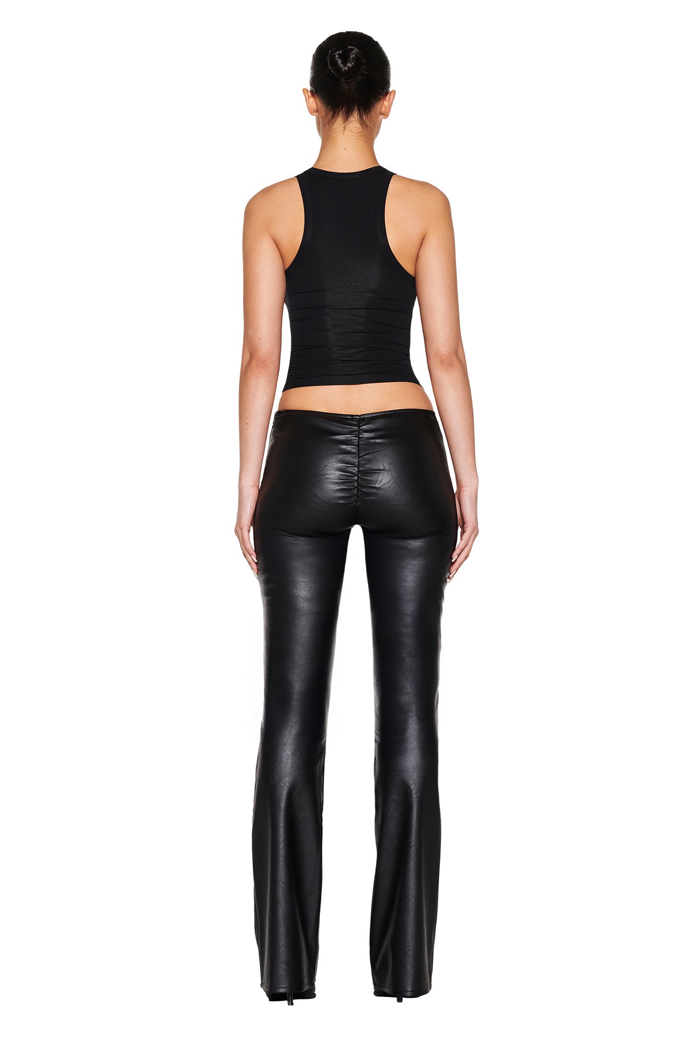 A person with their hair in a bun confidently poses against a plain white background, wearing the NEAVAH PU PANT - BLACK by I.AM.GIA and a black sleeveless top featuring sleek faux leather and a V-shaped waistband.