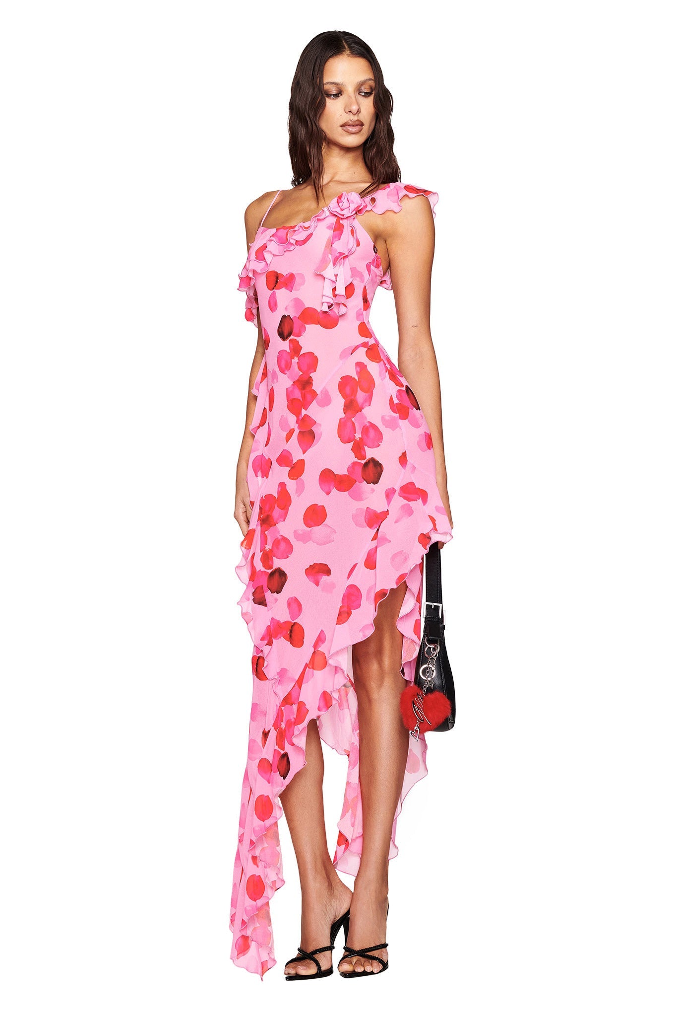 A woman showcases the I.AM.GIA JULIETTA MAXI DRESS in pink with a red polka dot pattern, ruffles, and an asymmetrical hem. She carries a black handbag and wears black high-heeled sandals against a white backdrop.