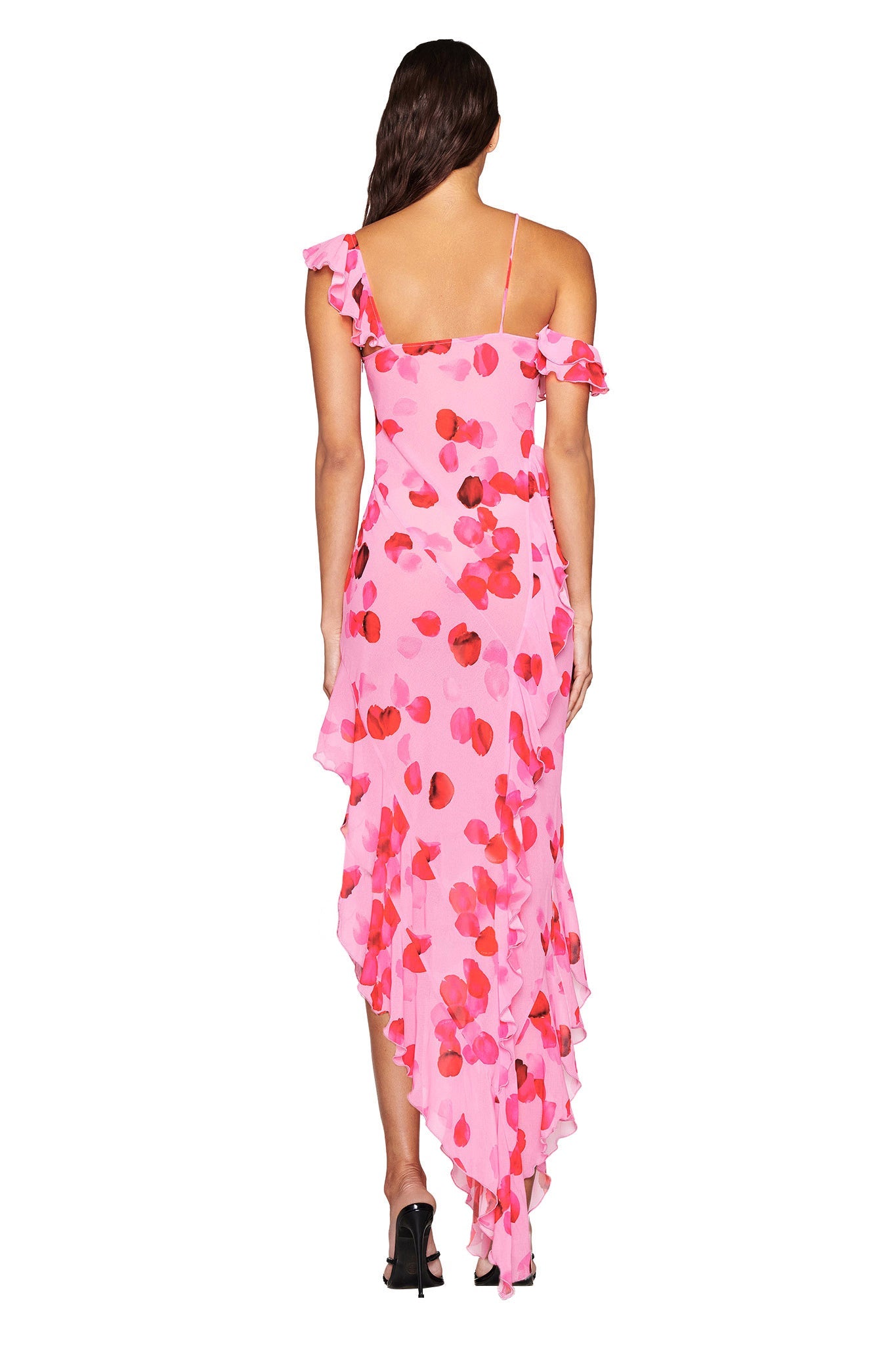 A woman in the JULIETTA MAXI DRESS - PINK by I.AM.GIA, with a PEACH PRETTY PETAL print, ruffled details, and an asymmetrical hem stands back to the camera. She completes her look with black high heels and long, wavy hair.