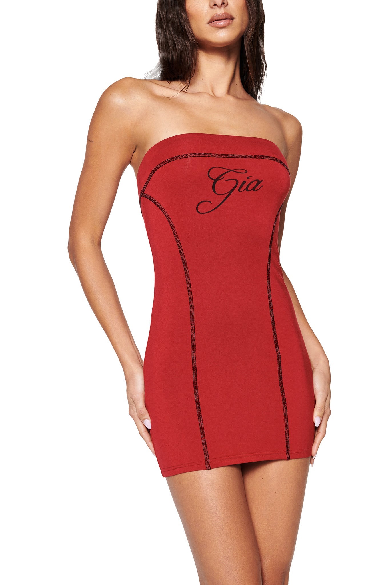 A woman confidently poses in the BLARE TUBE MINI DRESS - RED by I.AM.GIA, featuring contrast stitching and "Gia" boldly printed on the front. Her hands on her hips accentuate its bodycon fit against a plain white backdrop.