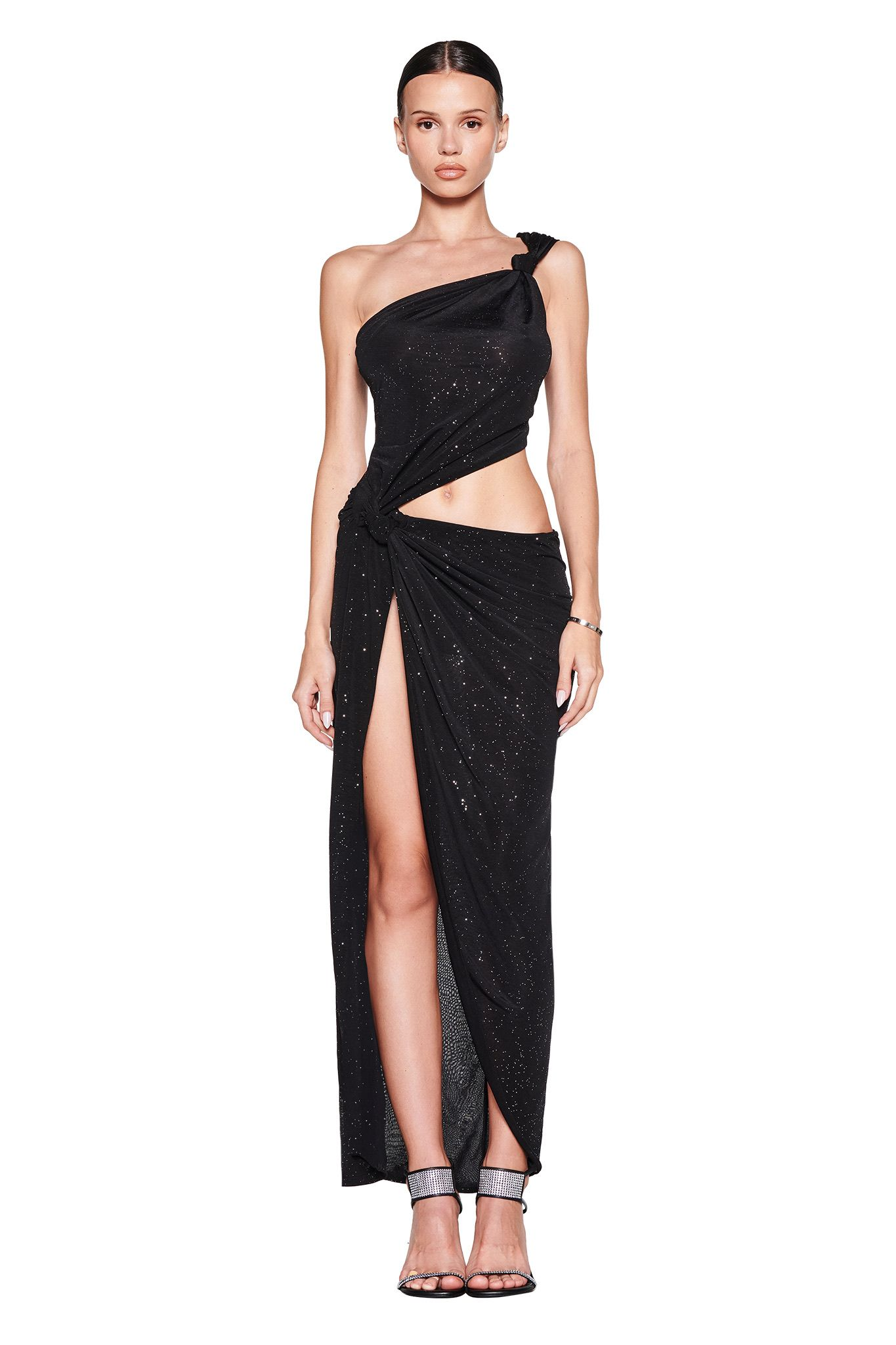 A woman faces forward in the I.AM.GIA ALIX MAXI DRESS - BLACK, a one-shoulder, high-slit dress with silver sparkles and waist cutout, paired with high-heeled sandals. Her straight hair is pulled back and she has a neutral expression.
