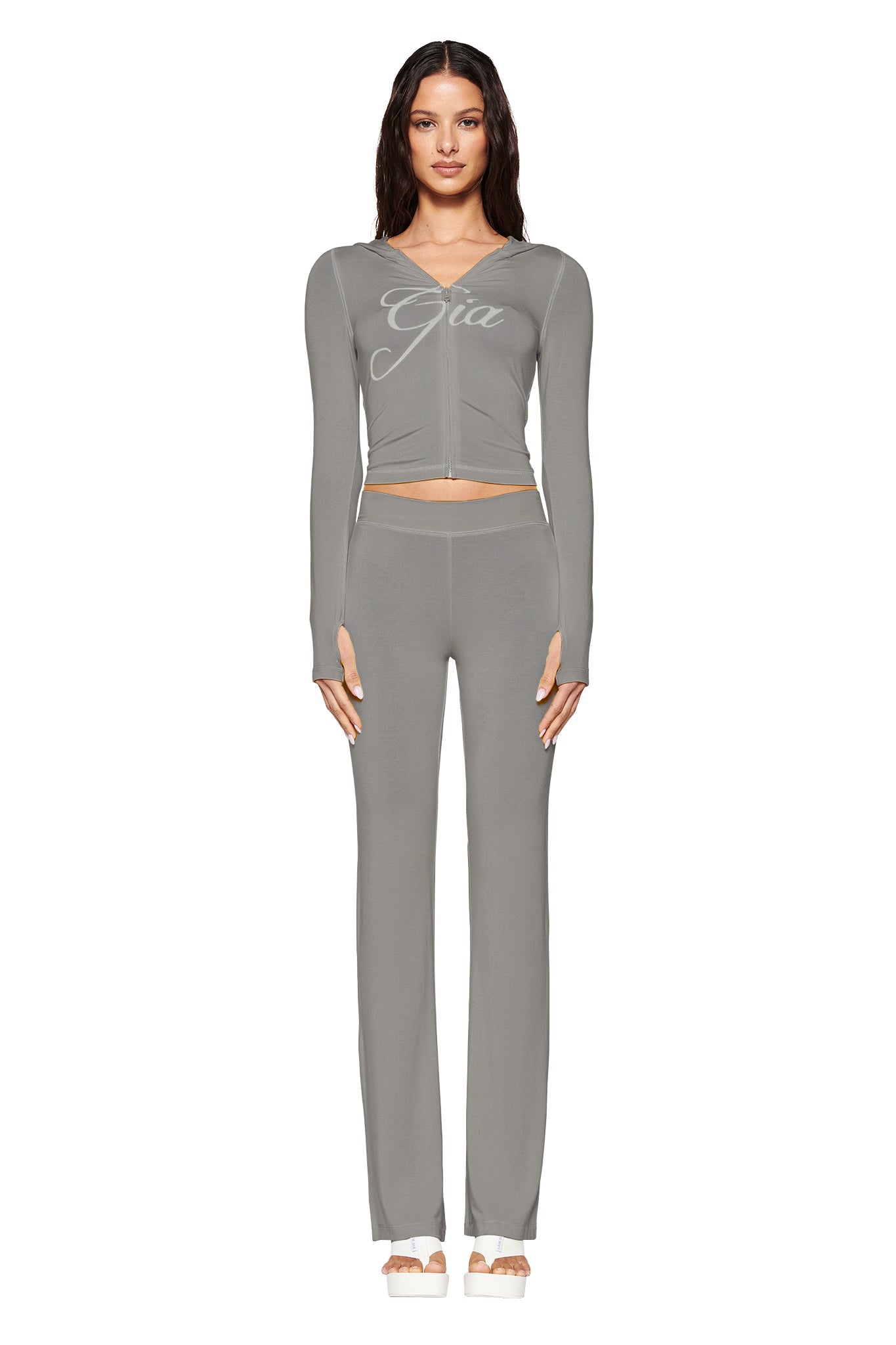 A person stands in a gray matching set with the I.AM.GIA Blare Hoodie in sage, featuring "Gia" on the top and high-waisted pants. They wear open-toed heels with long, wavy hair cascading down, and the outfit is crafted from bamboo fabric.