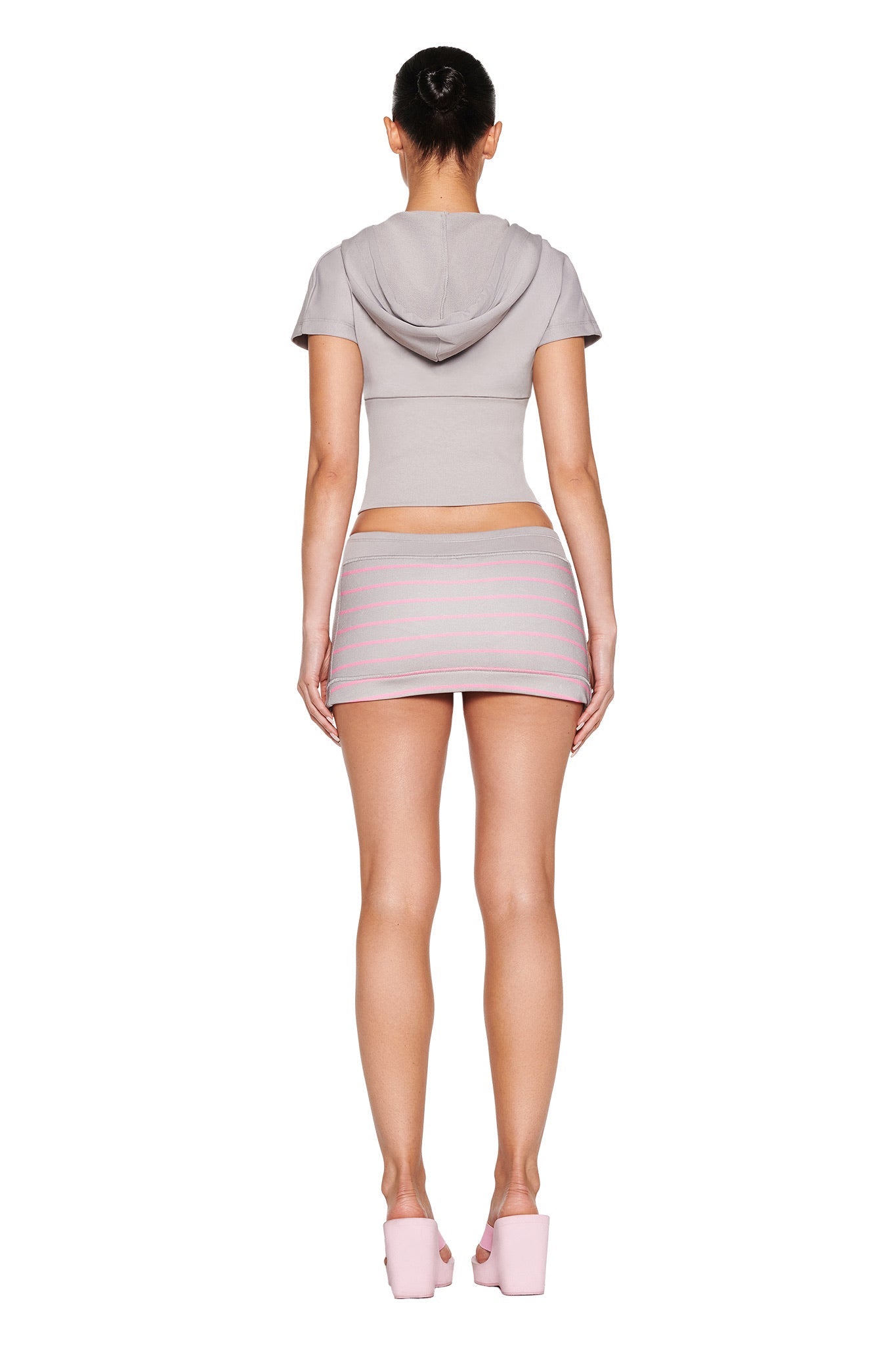 Rear view of a person in a short-sleeved cropped hoodie and I.AM.GIA's EZORA SKORT 2.0 - GREY : PINK with pastel stripes, showcasing a low rise waistline. Their hair is styled in a bun, paired with pink platform heels. The background is white.