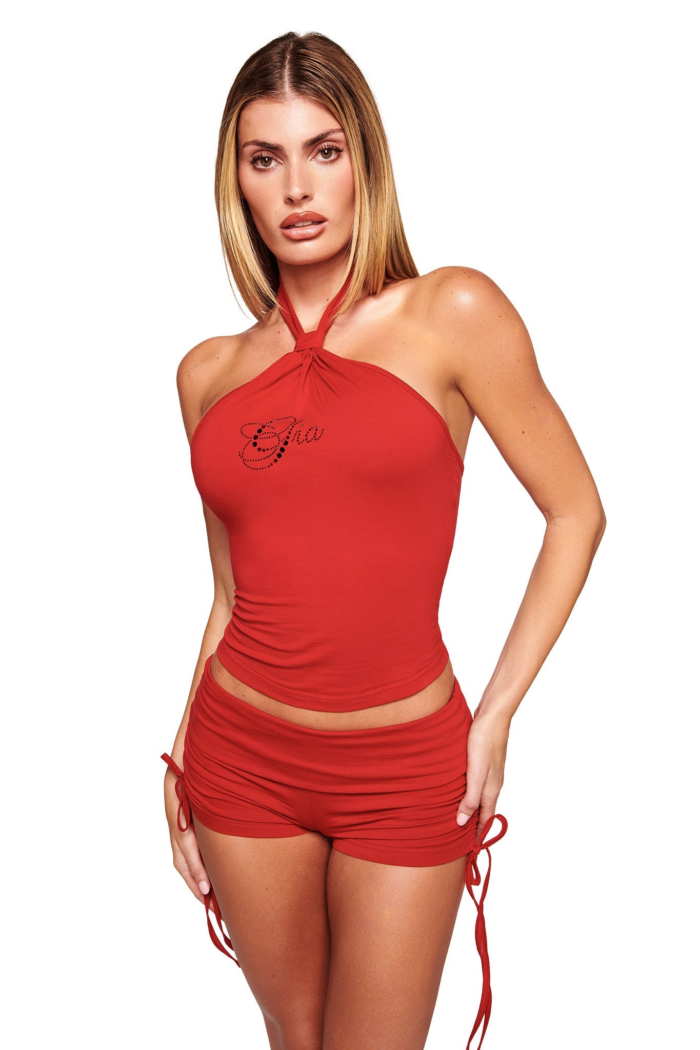 A woman with straight blonde hair wears the I.AM.GIA ROWAN TOP in rich red, a halter-neck style with diamonte logo detail, paired with matching red ruched bodycon shorts, standing against a white background.
