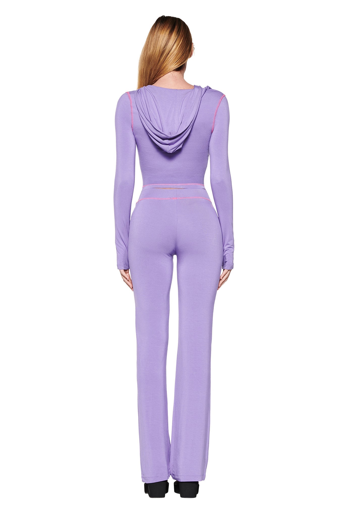 A woman with long hair is wearing the BLARE HOODIE - PURPLE jumpsuit by I.AM.GIA. It features a bodycon fit, long sleeves, and flared legs against a plain white background. She stands facing away.