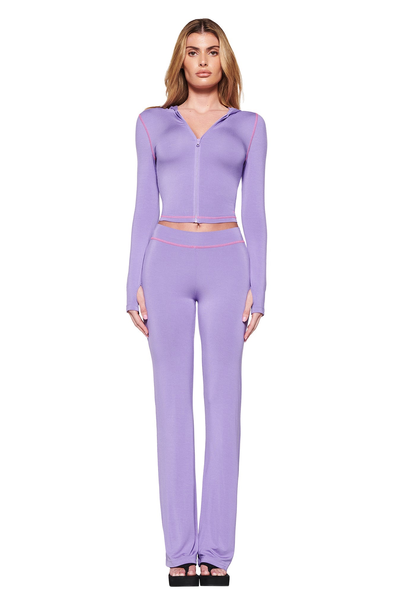 A woman models the I.AM.GIA BLARE HOODIE in purple, a fitted bodycon ensemble consisting of a long-sleeved zipper top and matching high-waisted pants made from stretchy bamboo. She poses against a white background in black platform shoes.