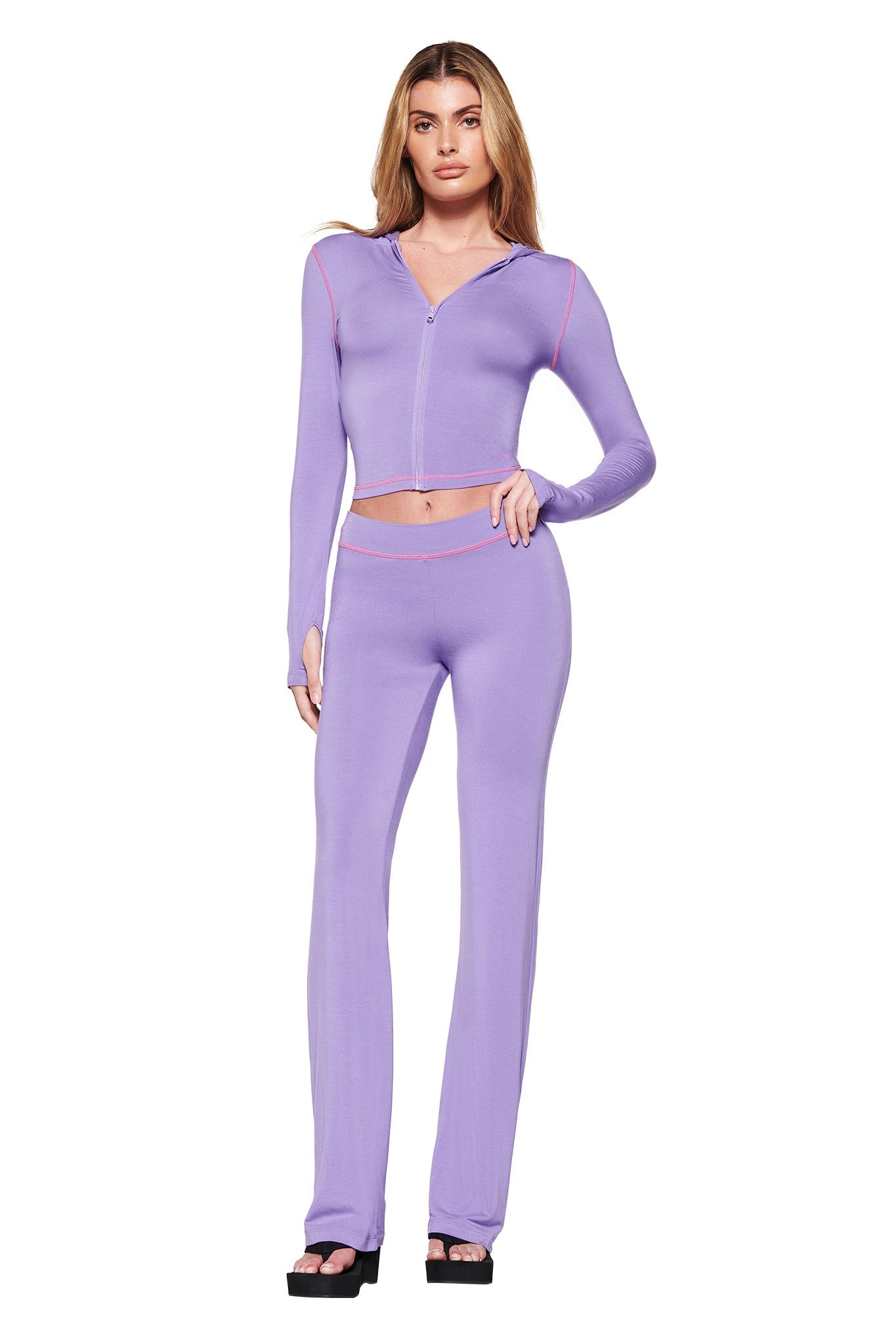 A woman poses in a fitted lavender hoodie and matching I.AM.GIA BLARE TRACKPANT - PURPLE with pink piping, sporting black platform sandals. Her long hair enhances the outfit's flattering bodycon silhouette against a plain white background.