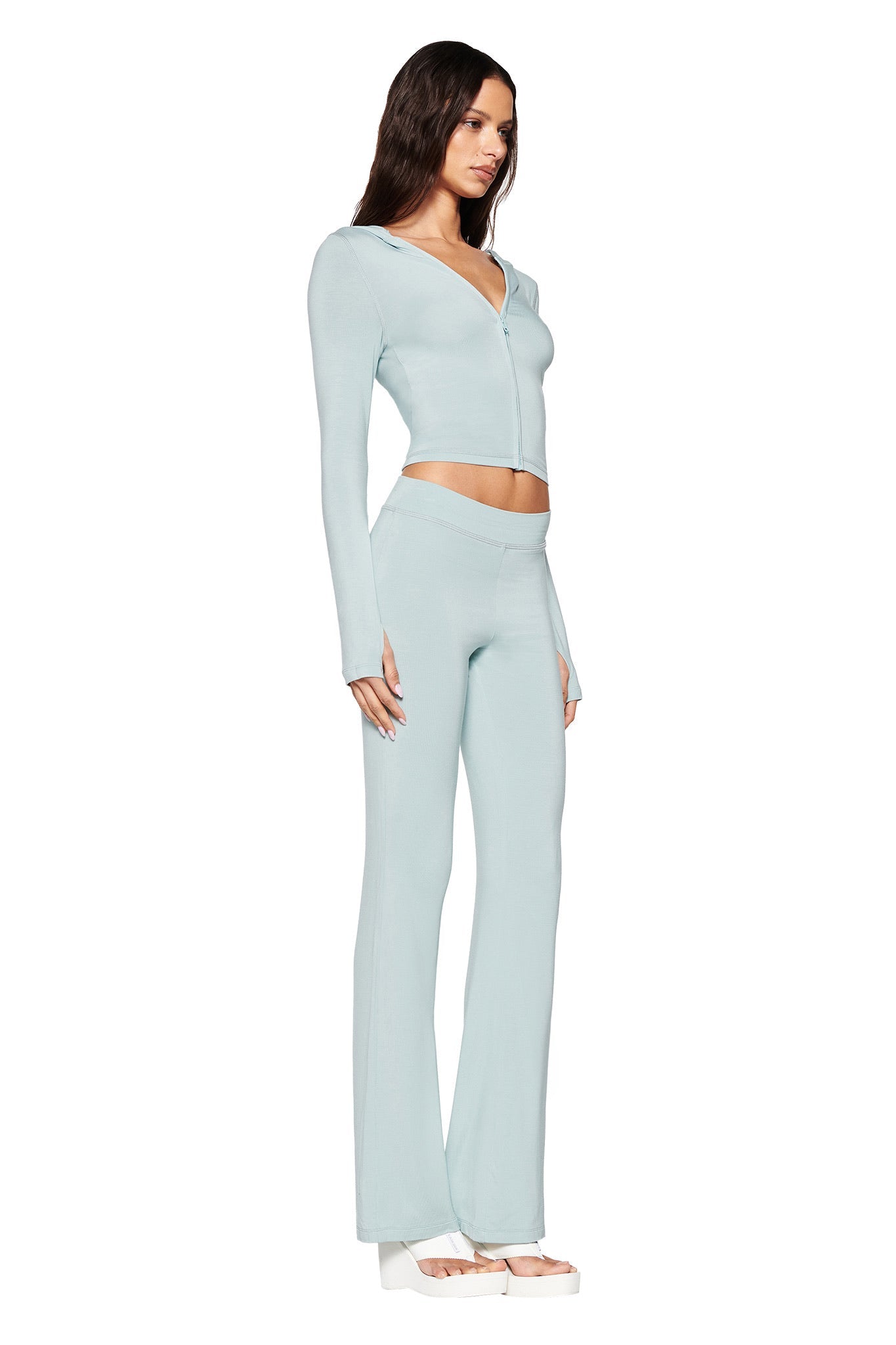 Against a minimalist white backdrop, a stylish individual stands in side profile, dressed in I.AM.GIA's seafoam long-sleeved cropped top with a zip front and the matching high-waisted BLARE TRACKPANT. They have long dark hair and are wearing white platform sandals.