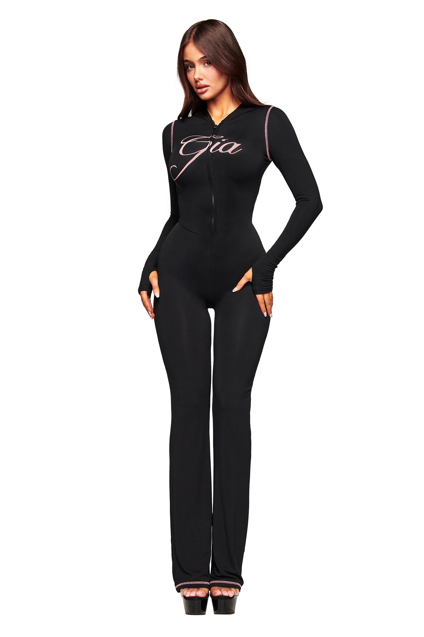 A woman with long brown hair faces forward in the fitted black BLARE JUMPSUIT by I.AM.GIA, featuring long sleeves, "Gia" script on the chest, and flared legs. She wears black platform heels and stands against a white background.