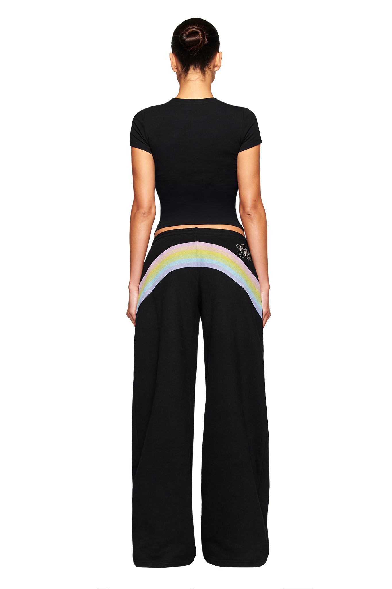 A woman with her hair in a neat bun faces away, wearing the I.AM.GIA FAFI T-SHIRT in black and matching wide-leg pants with a rainbow design on the back, set against a plain white background.