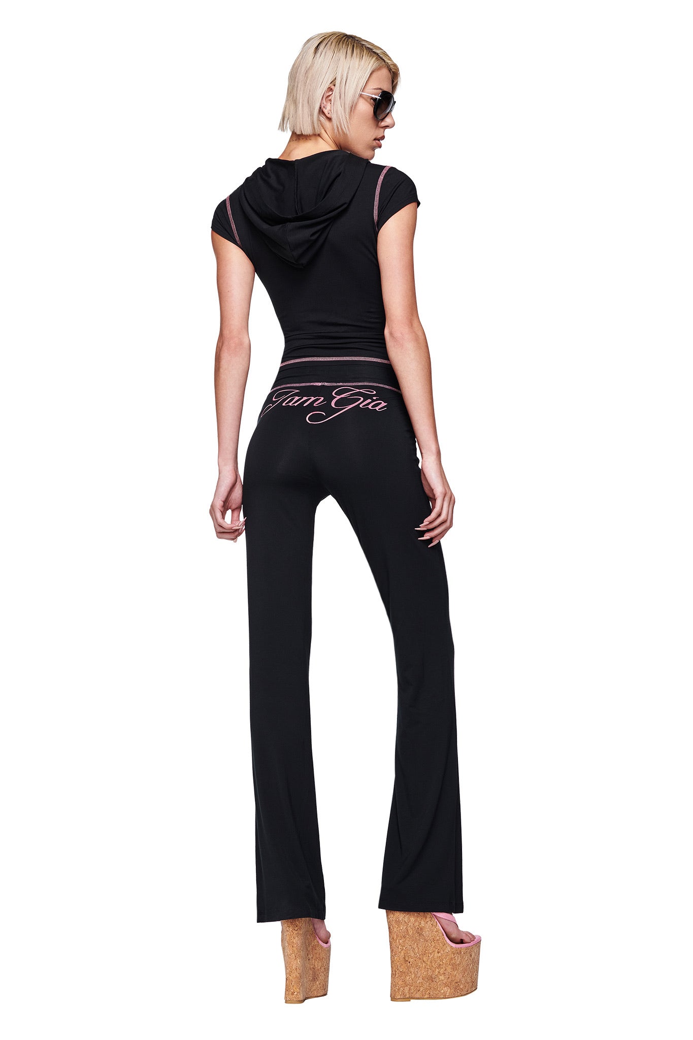 Wearing the I.AM.GIA BLARE CAP SLEEVE HOODIE in black and sunglasses, a person with short blonde hair poses backward in black flared I.AM.GIA pants featuring "VAN GIA" script on the back, paired with tall cork platform shoes.