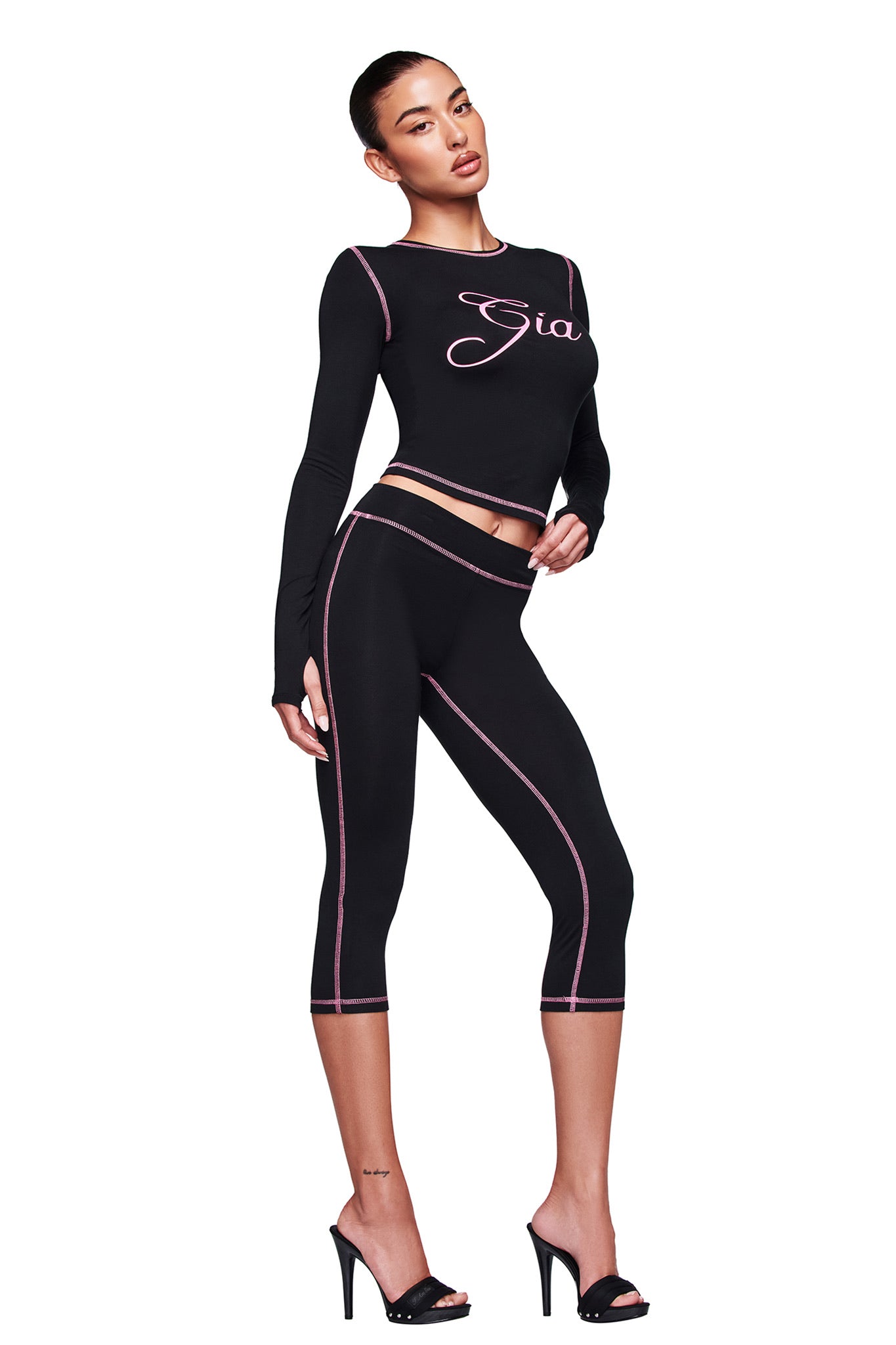 A woman poses in the fitted black BLARE LONG SLEEVE TOP by I.AM.GIA with pink stitching and “Gia” text, paired with matching cropped leggings and black heels, against a white background.