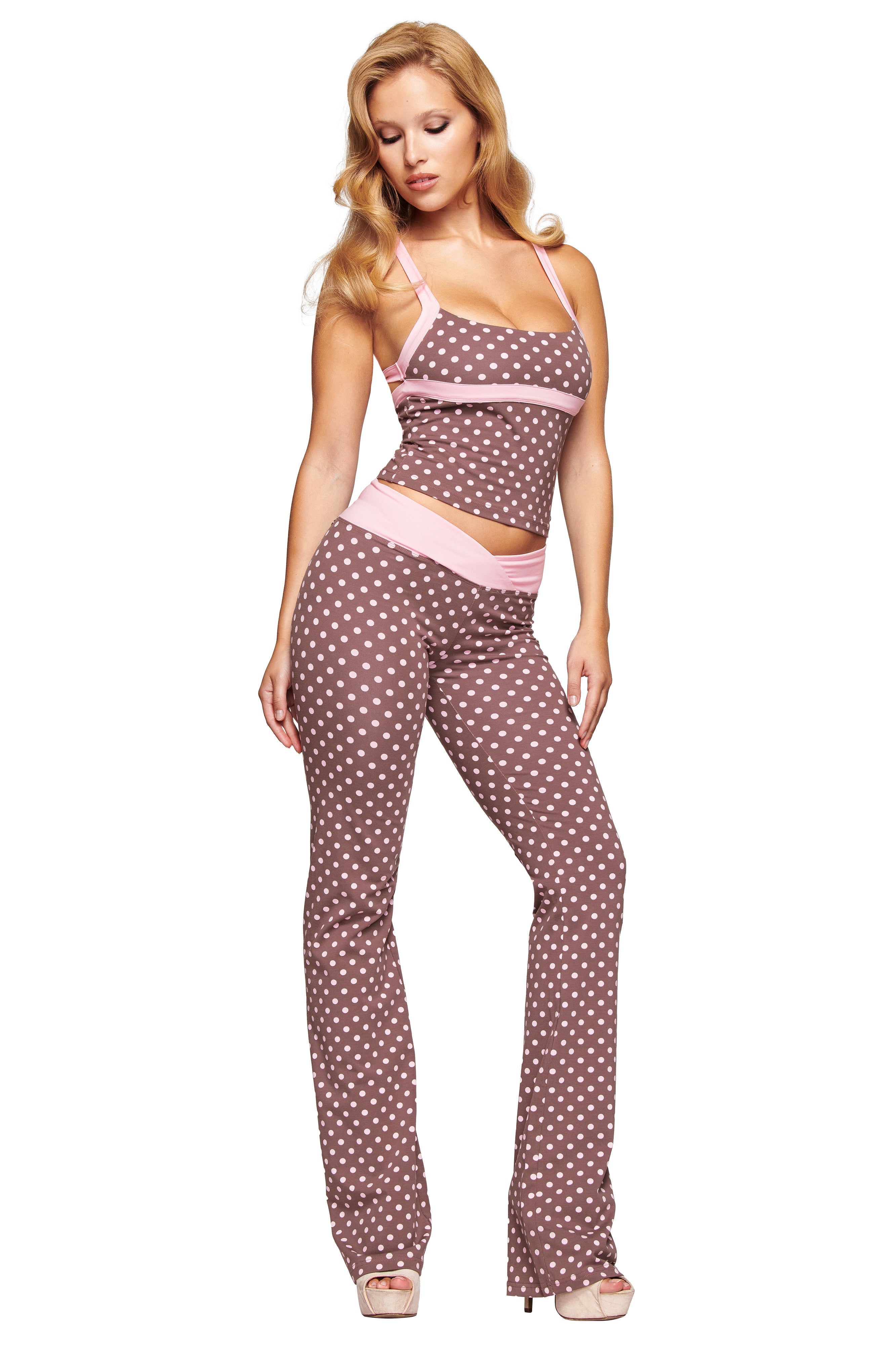 A woman poses in a sleeveless, fitted brown top and I.AM.GIA BLARE CROSSOVER PANT - PINK CHOCOLATE with a mid rise waist, white polka dot print, light pink trim, and high-heeled shoes.