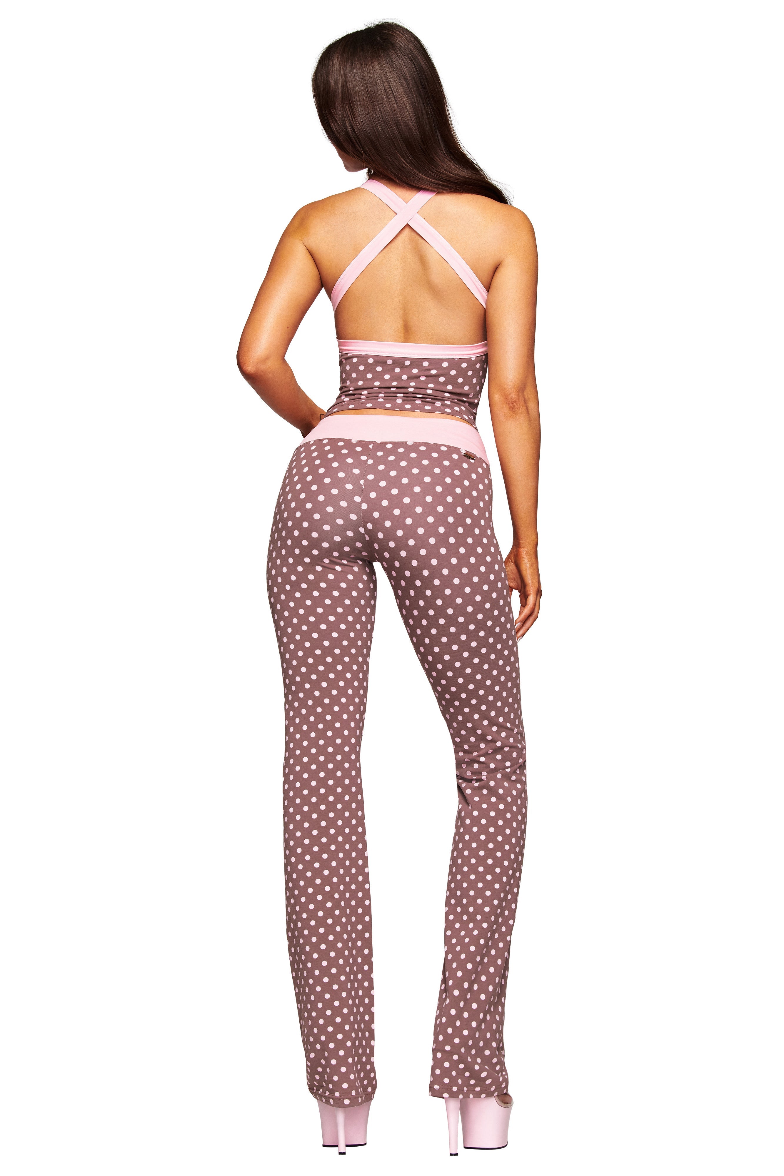 A woman faces away, wearing the I.AM.GIA BLARE CROSSOVER PANT - PINK CHOCOLATE with white polka dots and pink trim, a brown halter top, and light pink platform heels, set against a white background.