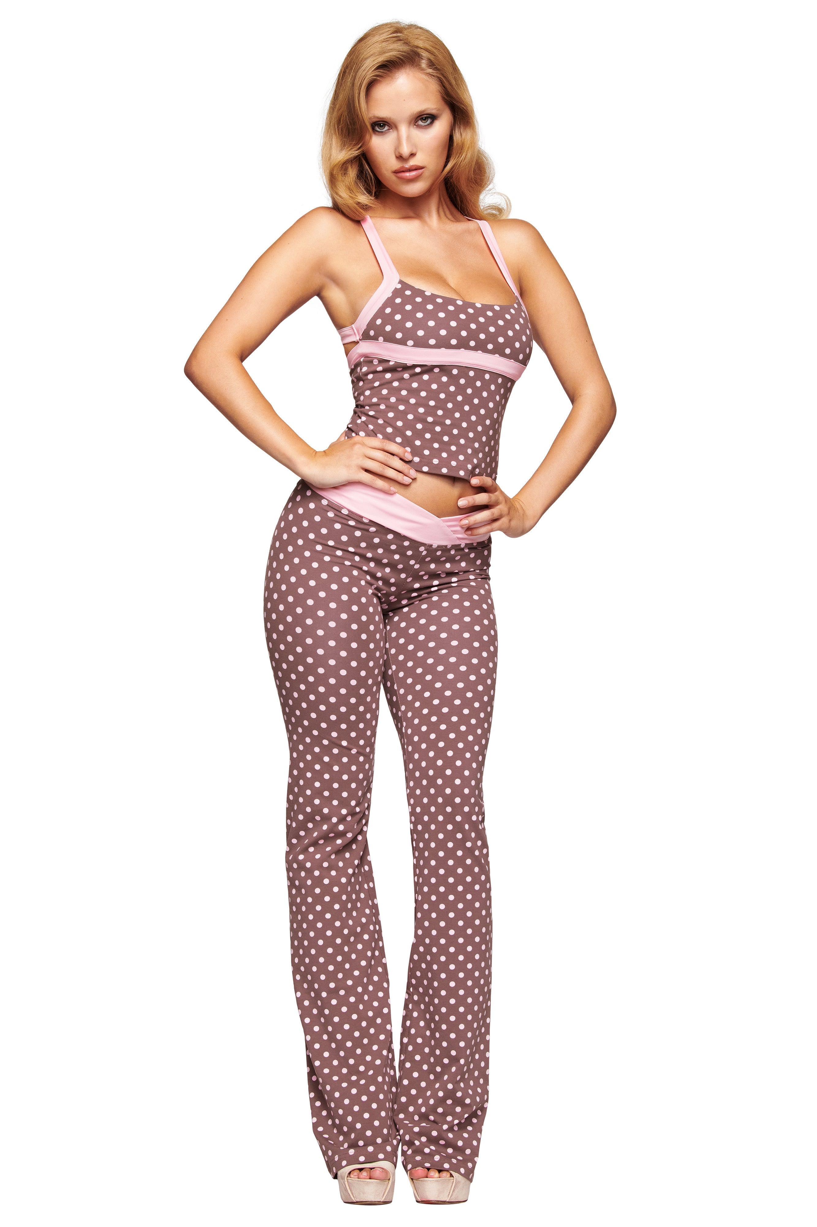 A woman poses in the I.AM.GIA BLARE CROSSOVER PANT - PINK CHOCOLATE, featuring a brown and pink polka dot print, paired with a matching sleeveless fitted top and light pink heels against a white background.